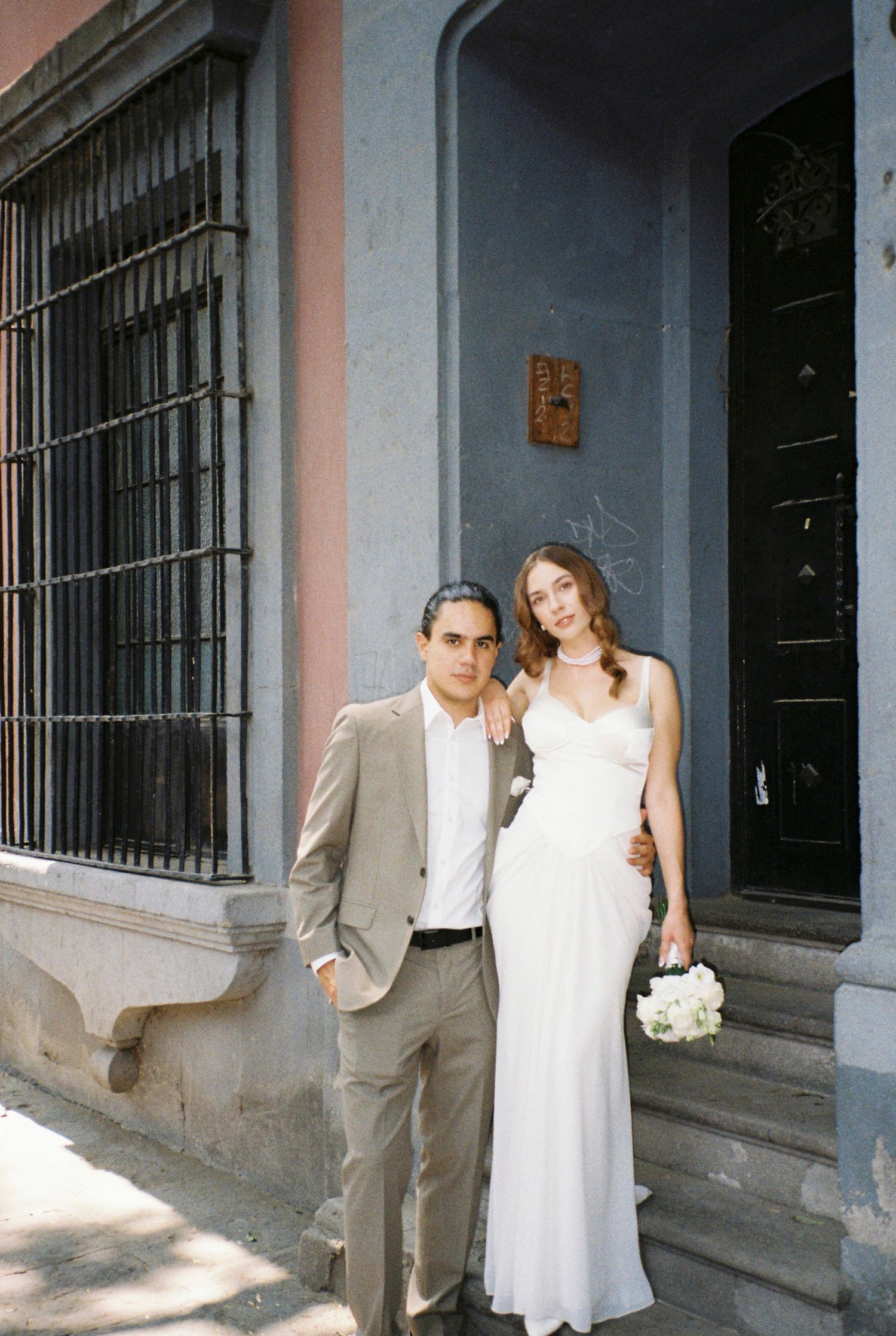 Couple posing together against a grey building wall with barred windows.