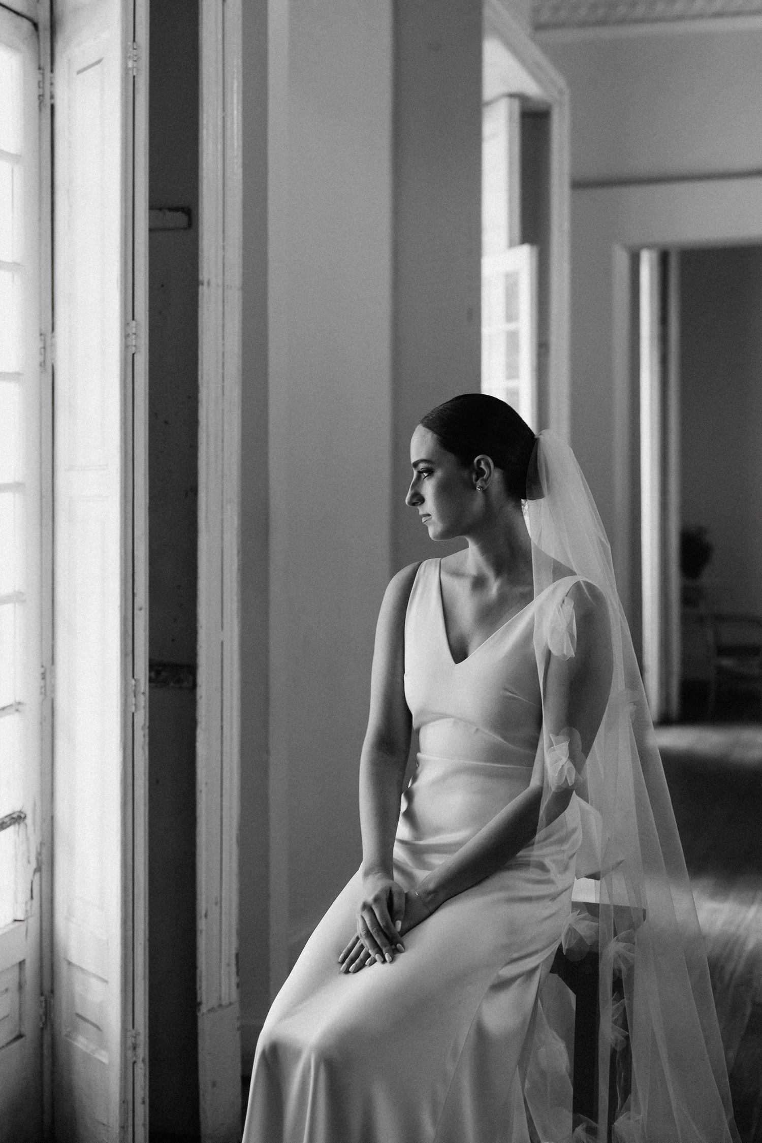 Elegant bride in minimalist white dress sits pensively by window in black and white photograph.