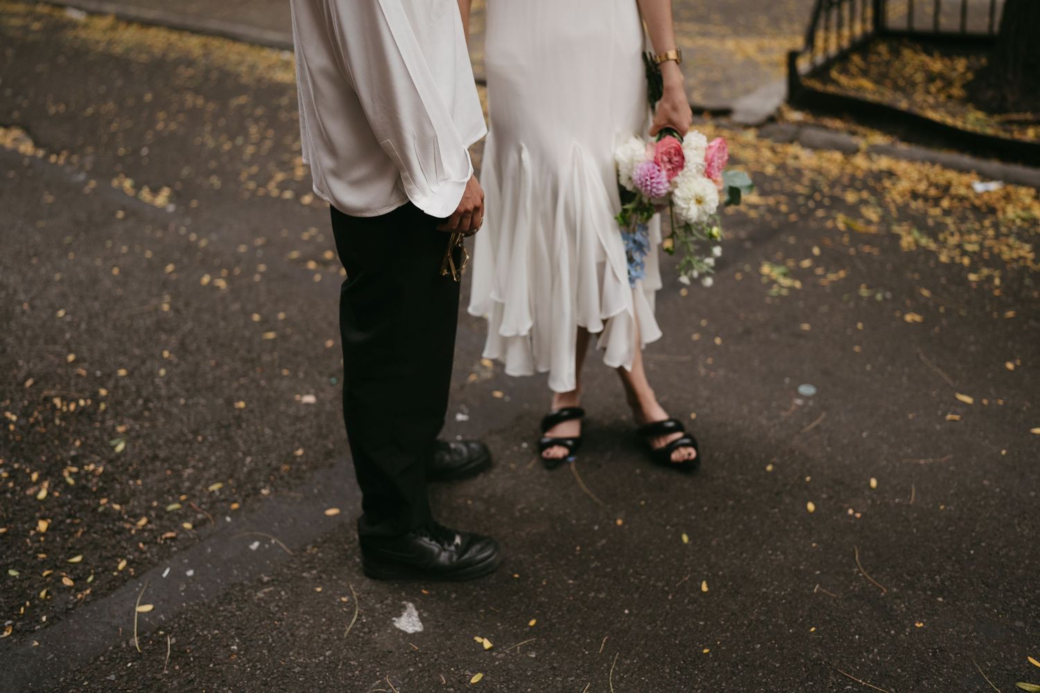A couple in formal attire stands together on a sidewalk with fallen autumn leaves.