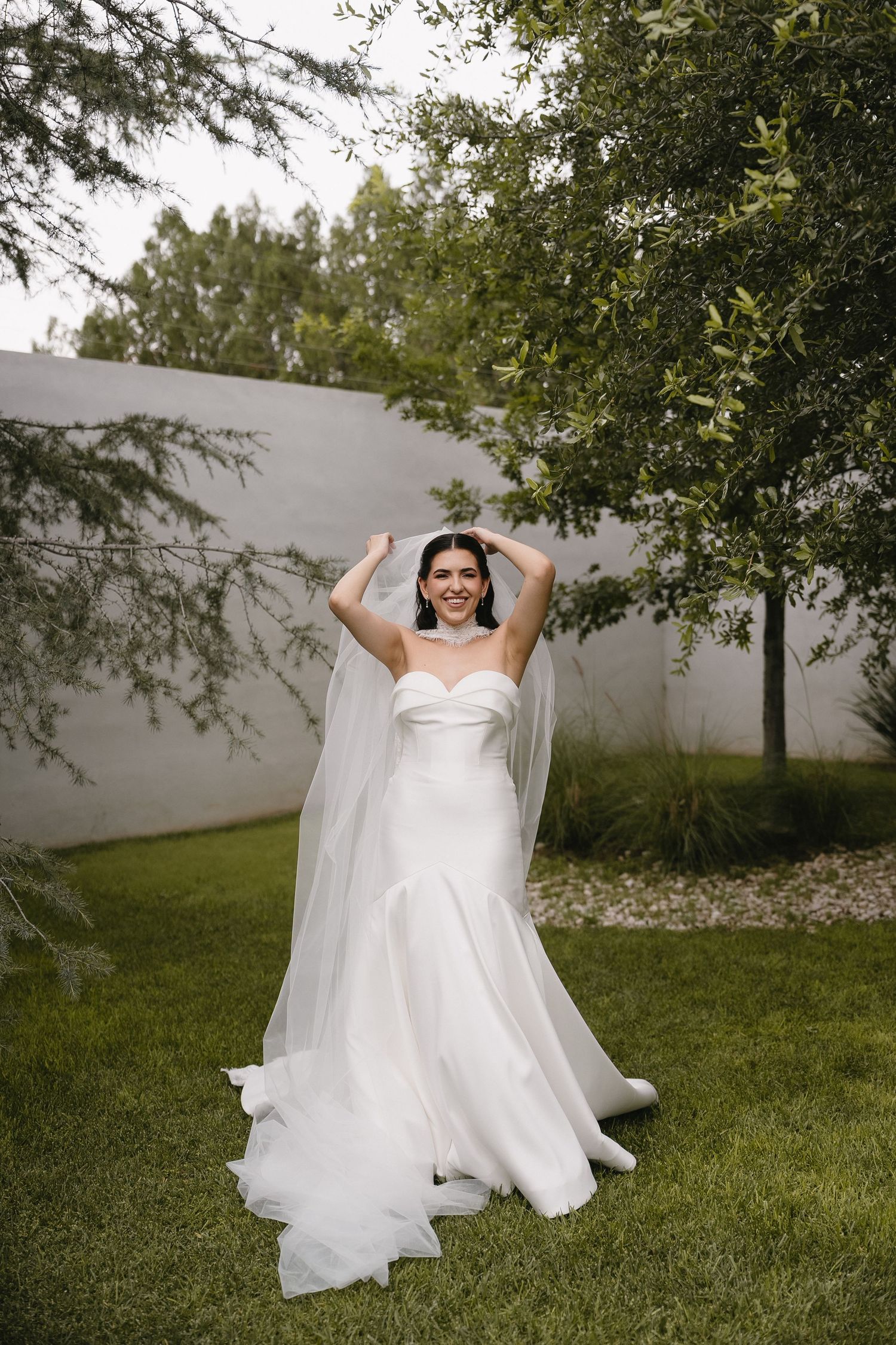 A bride in a flowing white wedding dress poses joyfully in a garden setting.