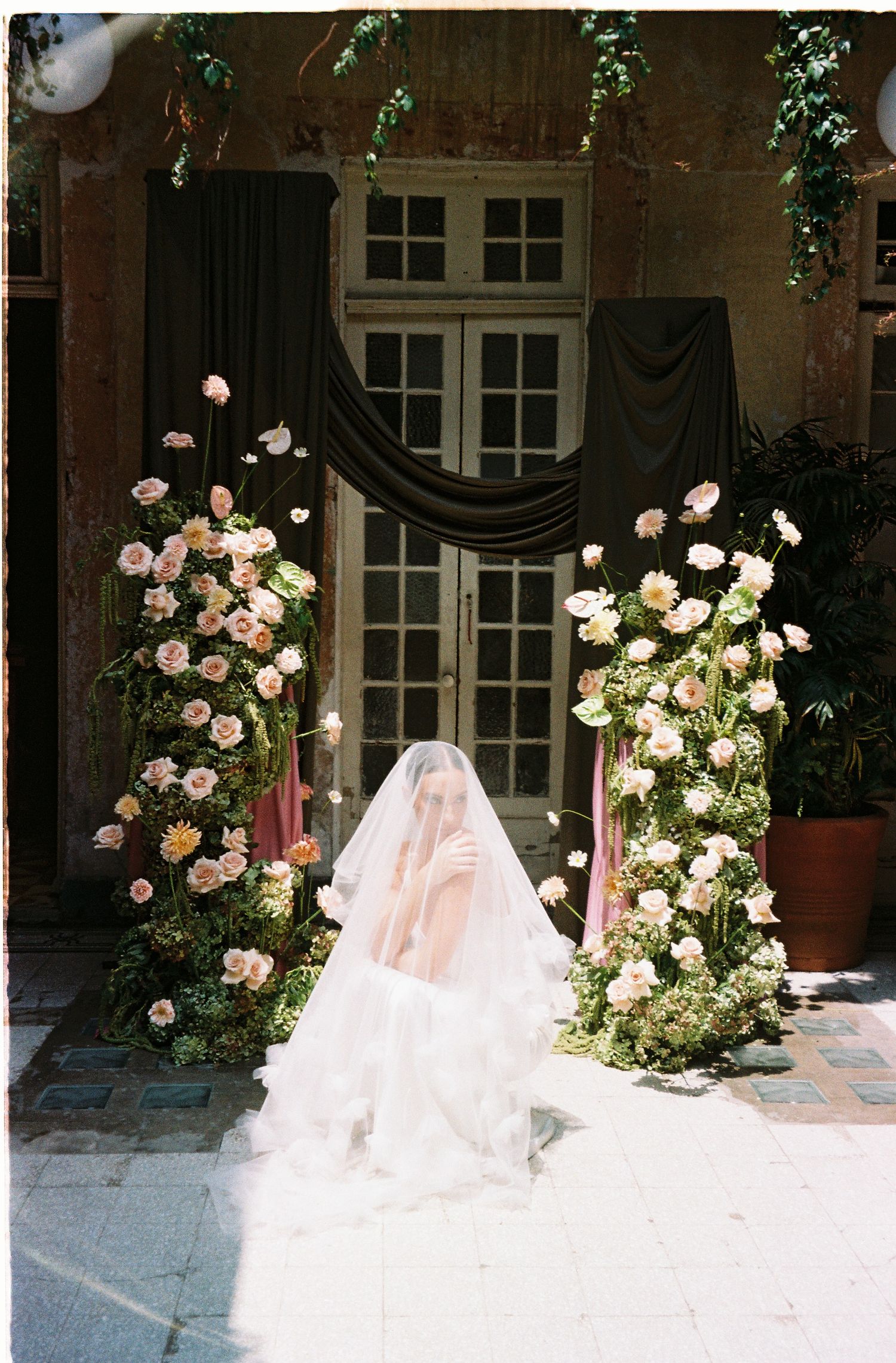 A flowing white wedding dress train trails elegantly across a stone courtyard surrounded by blooming rose bushes.
