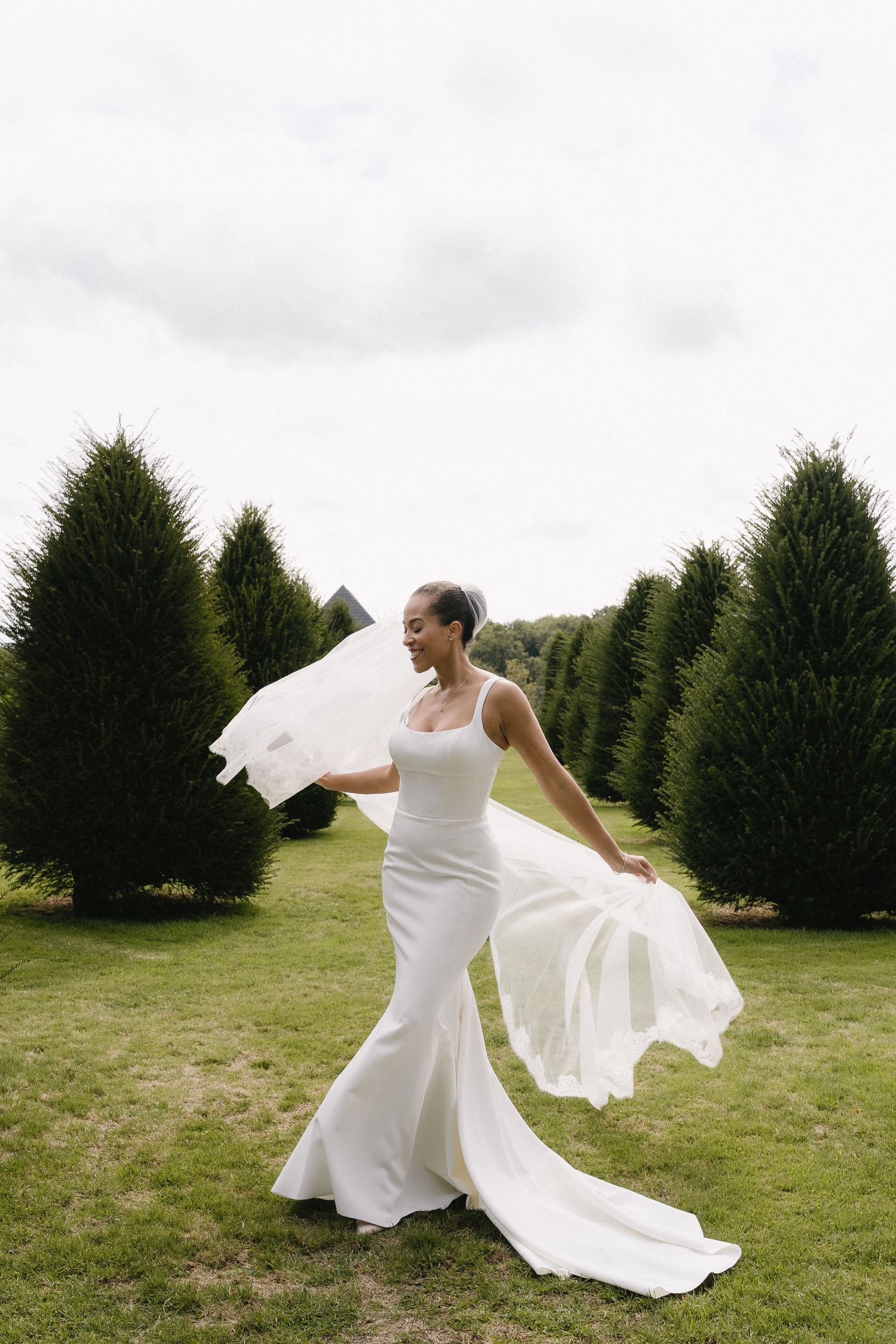 A bride in a fitted white dress twirling her veil in a garden surrounded by evergreen trees.