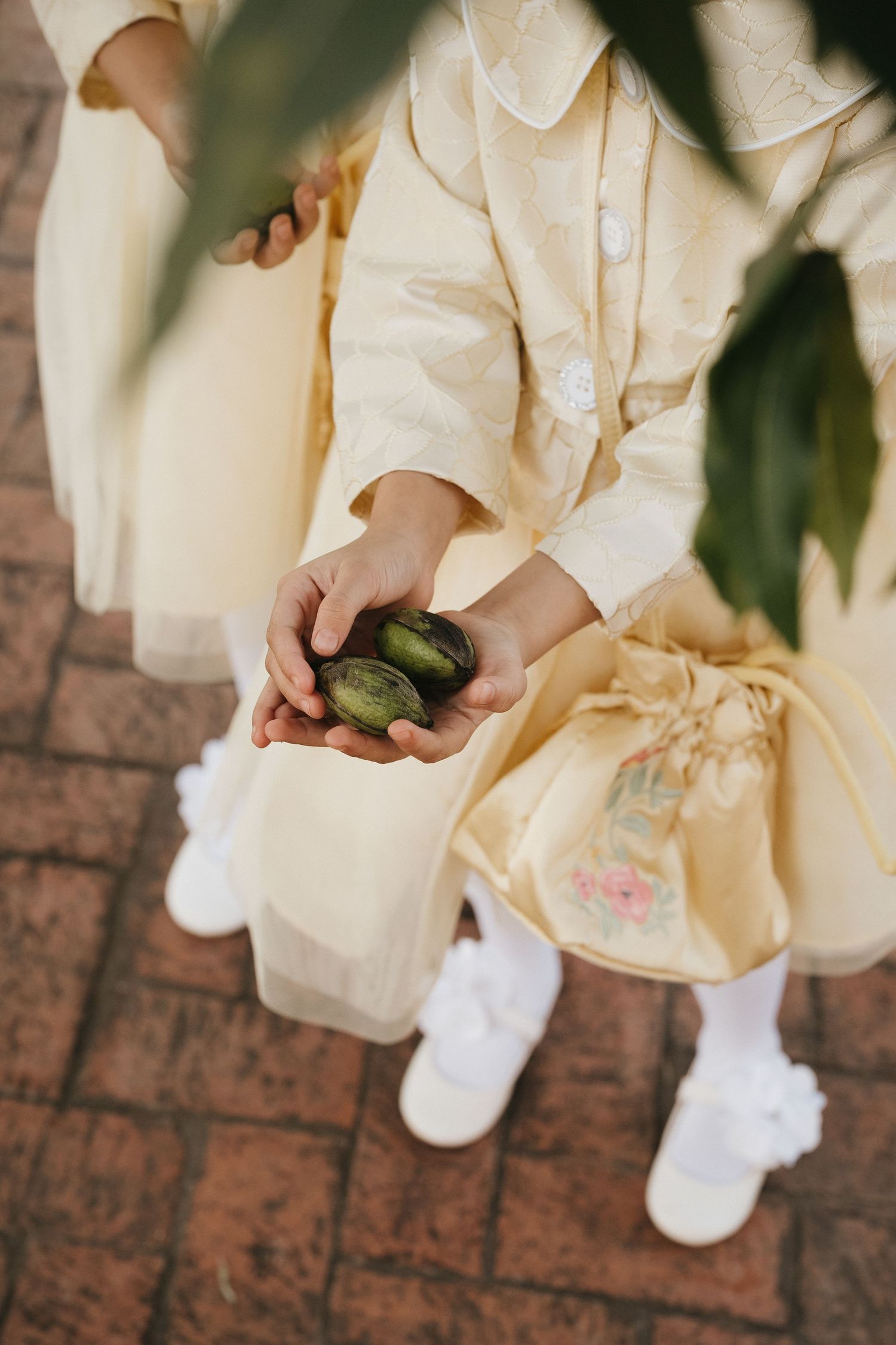 Person in cream-colored clothing holding green objects on brick pavement.