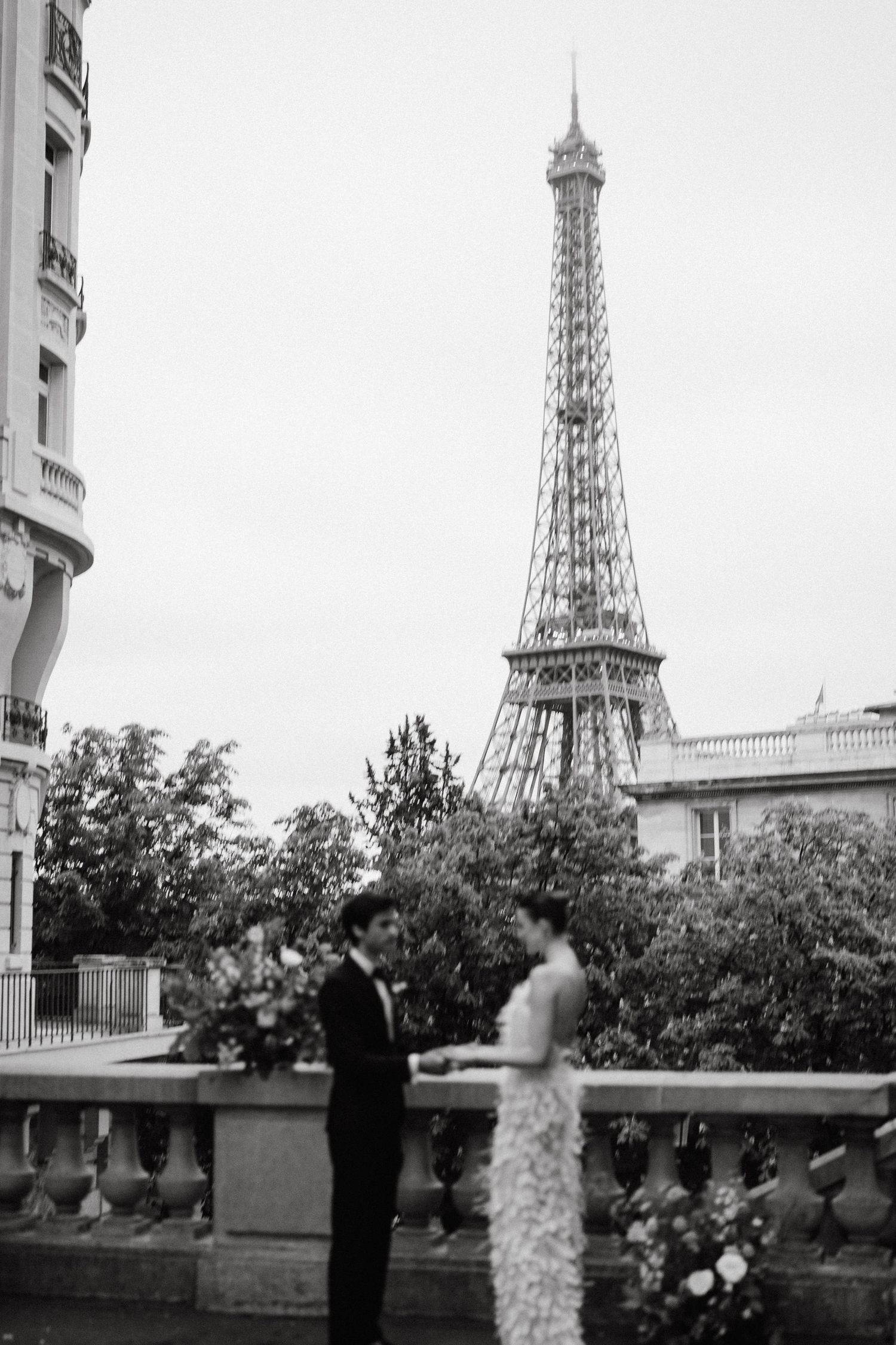 Black and white photograph of the Eiffel Tower visible through trees and buildings in Paris.