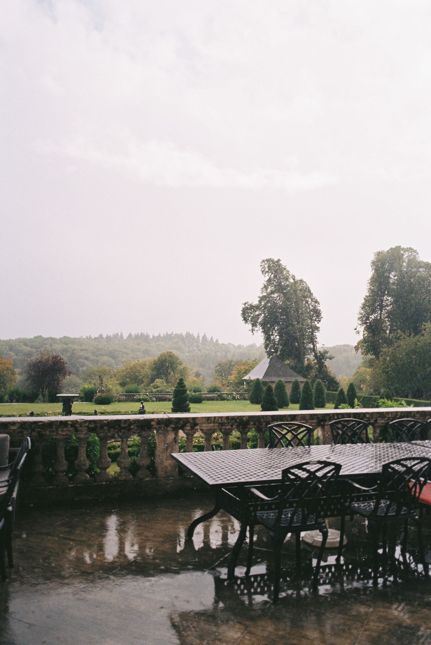 Panoramic view of a rain-soaked outdoor terrace with wrought iron furniture overlooking misty hills and trees.