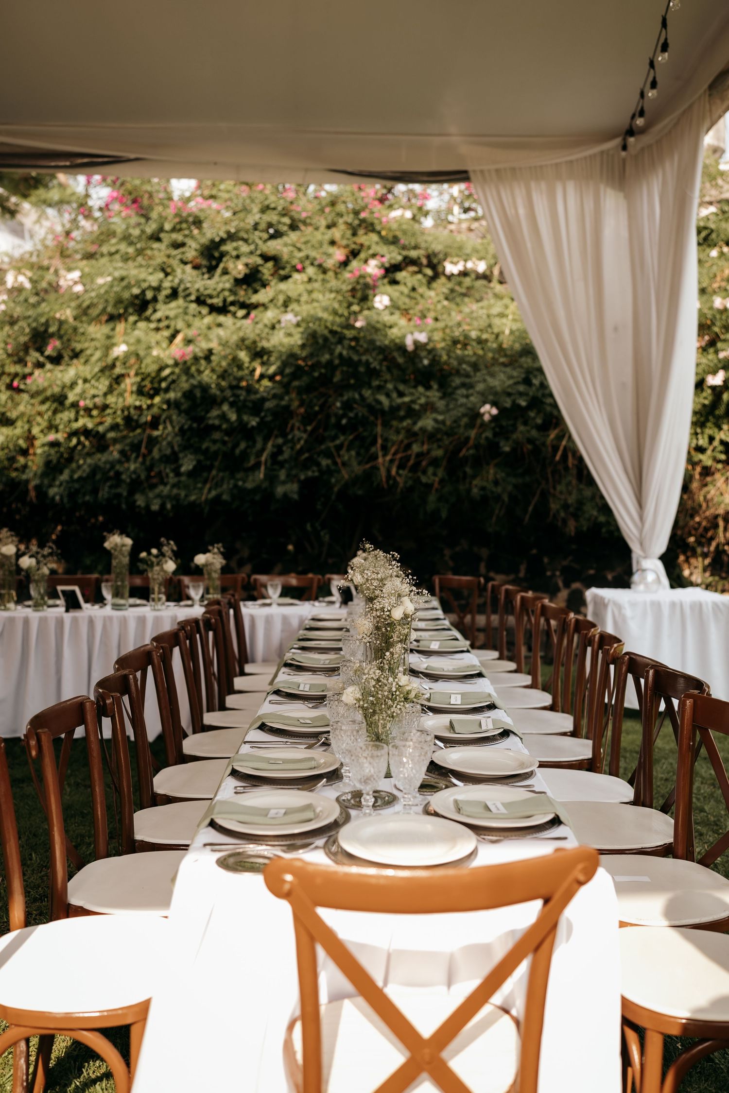 Elegant wedding reception setup with white tablecloths and wooden chairs under a draped tent.