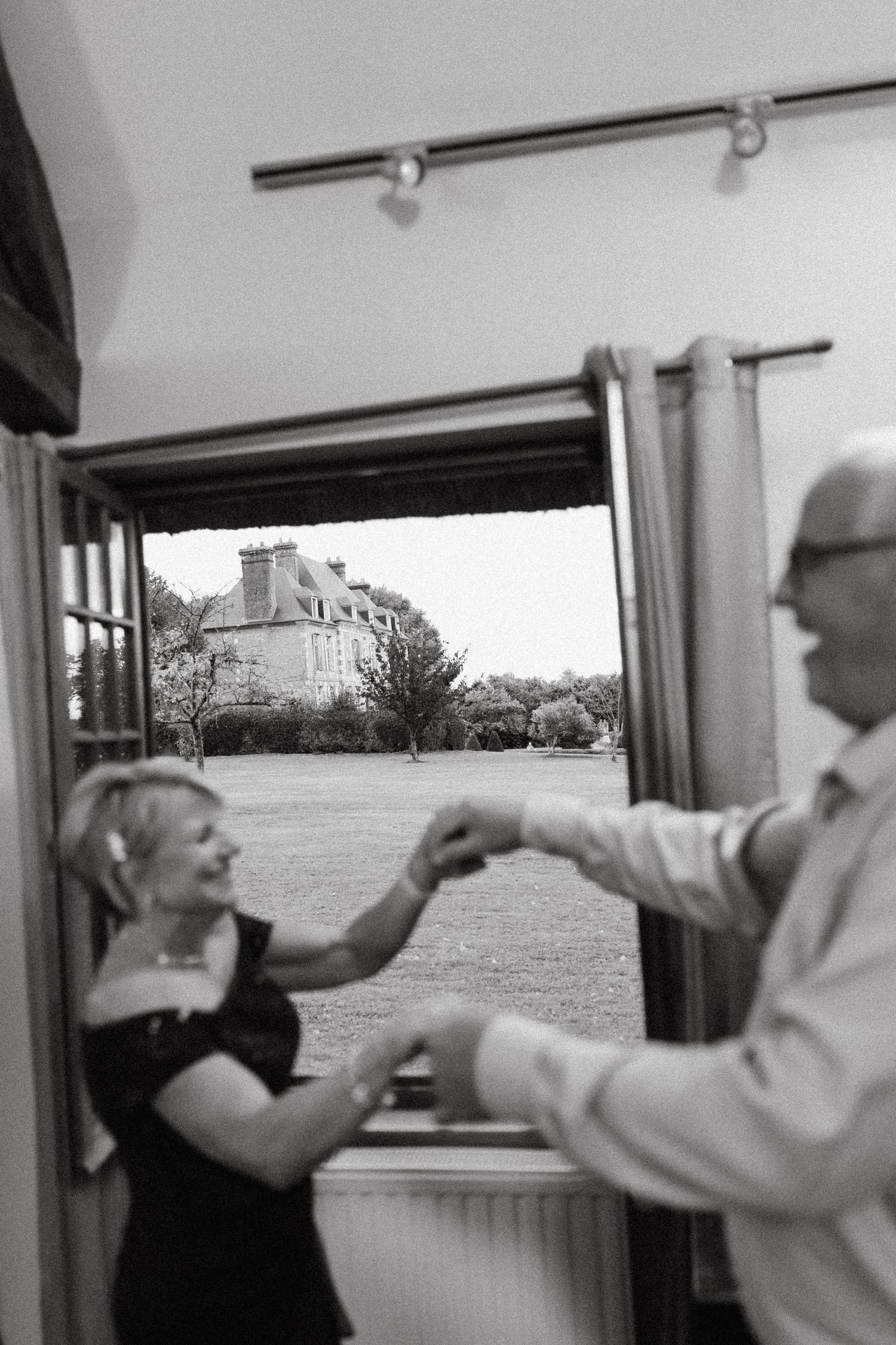 A black and white photo shows two people dancing together near a window with a view of a stately building outside.