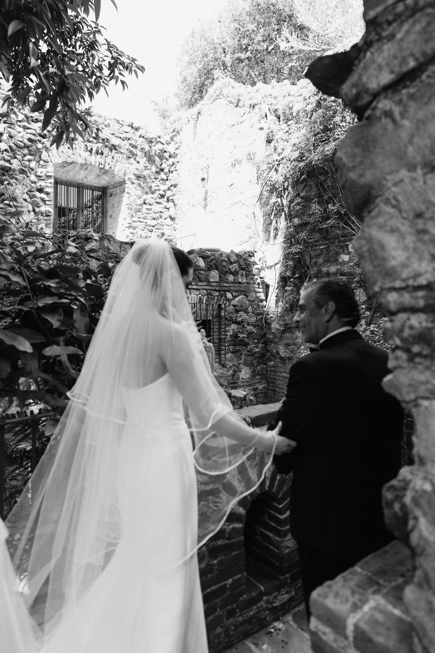 Wedding couple walking through stone archway in garden setting.