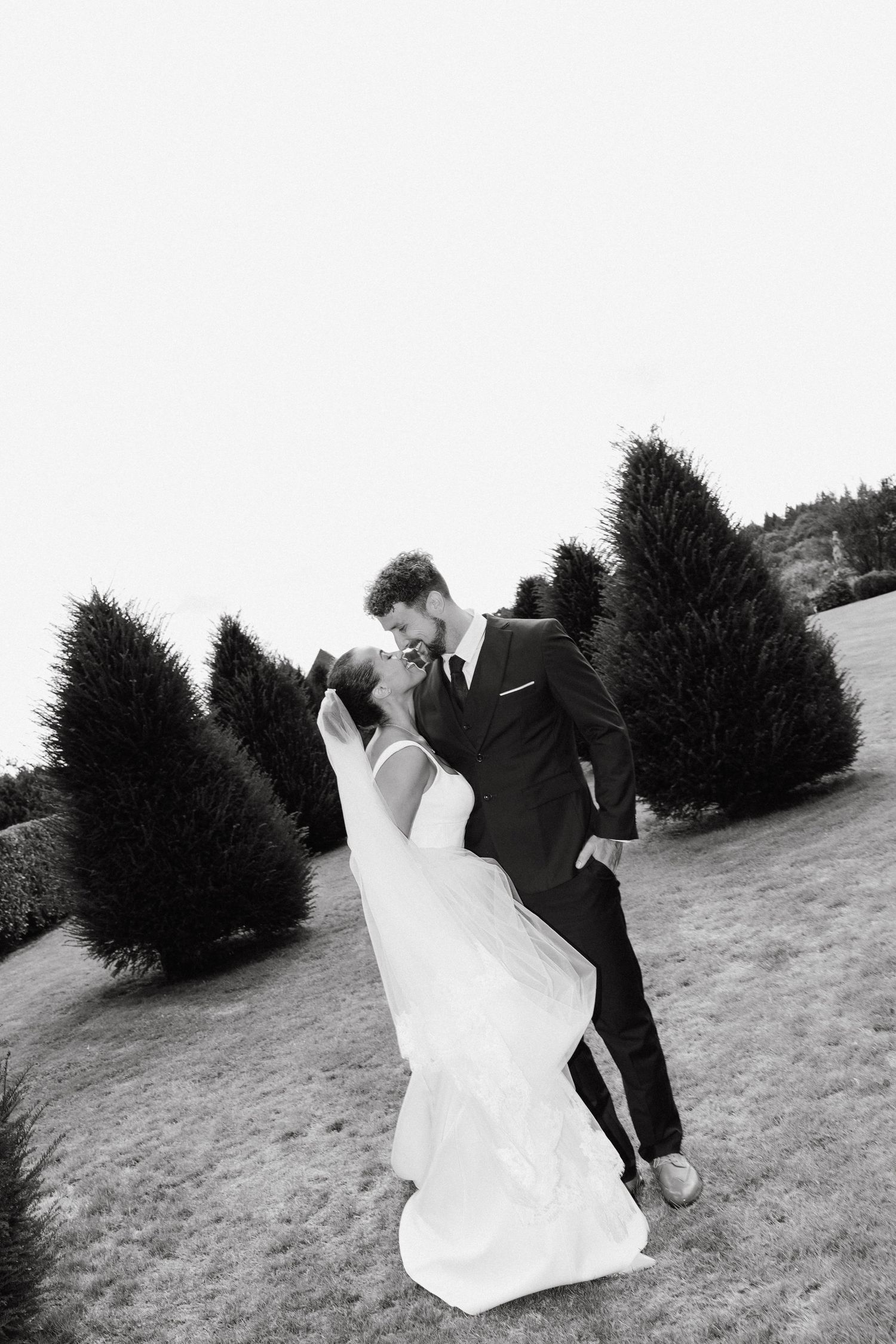 Black and white photo of bride and groom sharing a romantic kiss outdoors among evergreen trees on their wedding day.