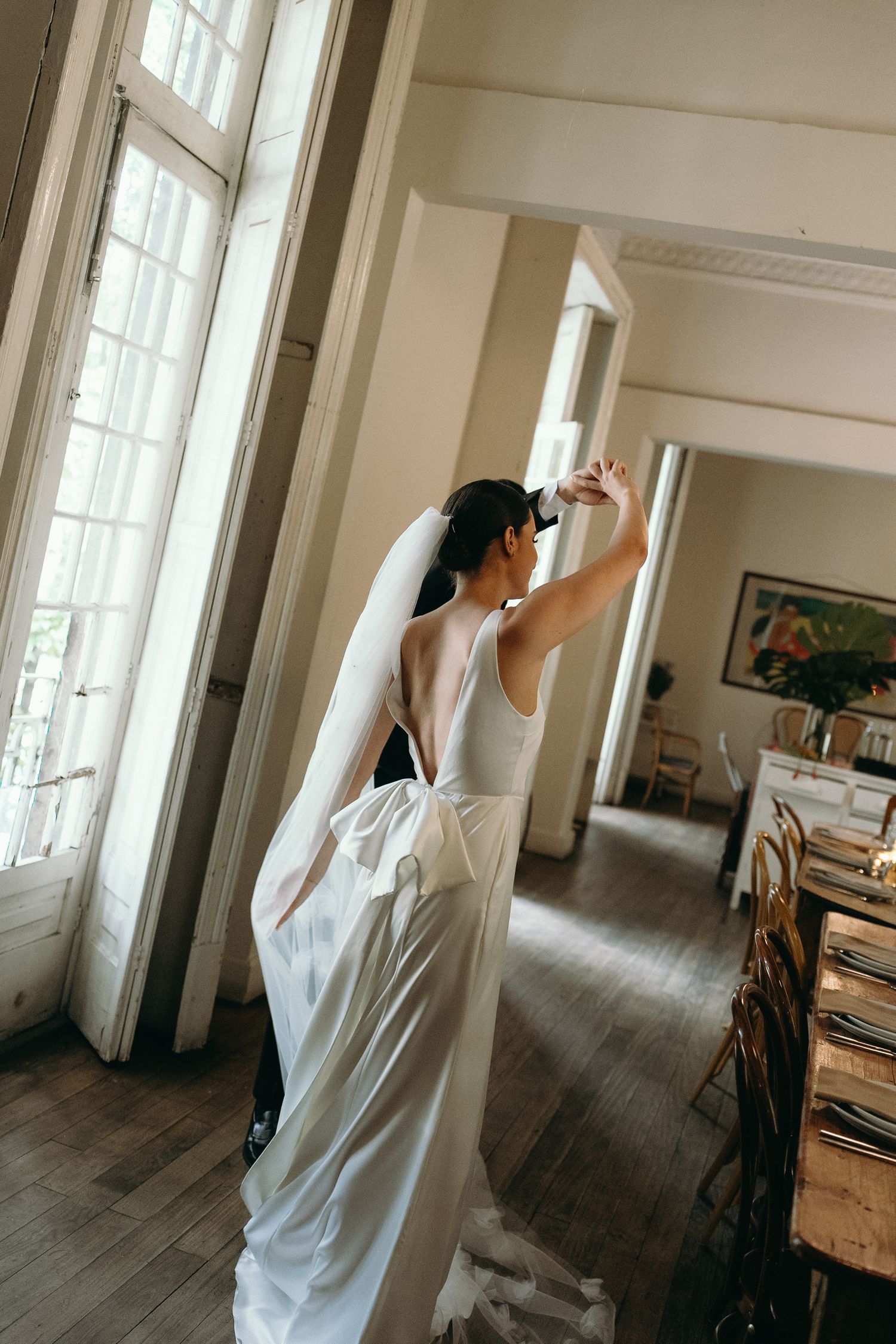 Bride in flowing white gown poses gracefully near tall windows in vintage interior.