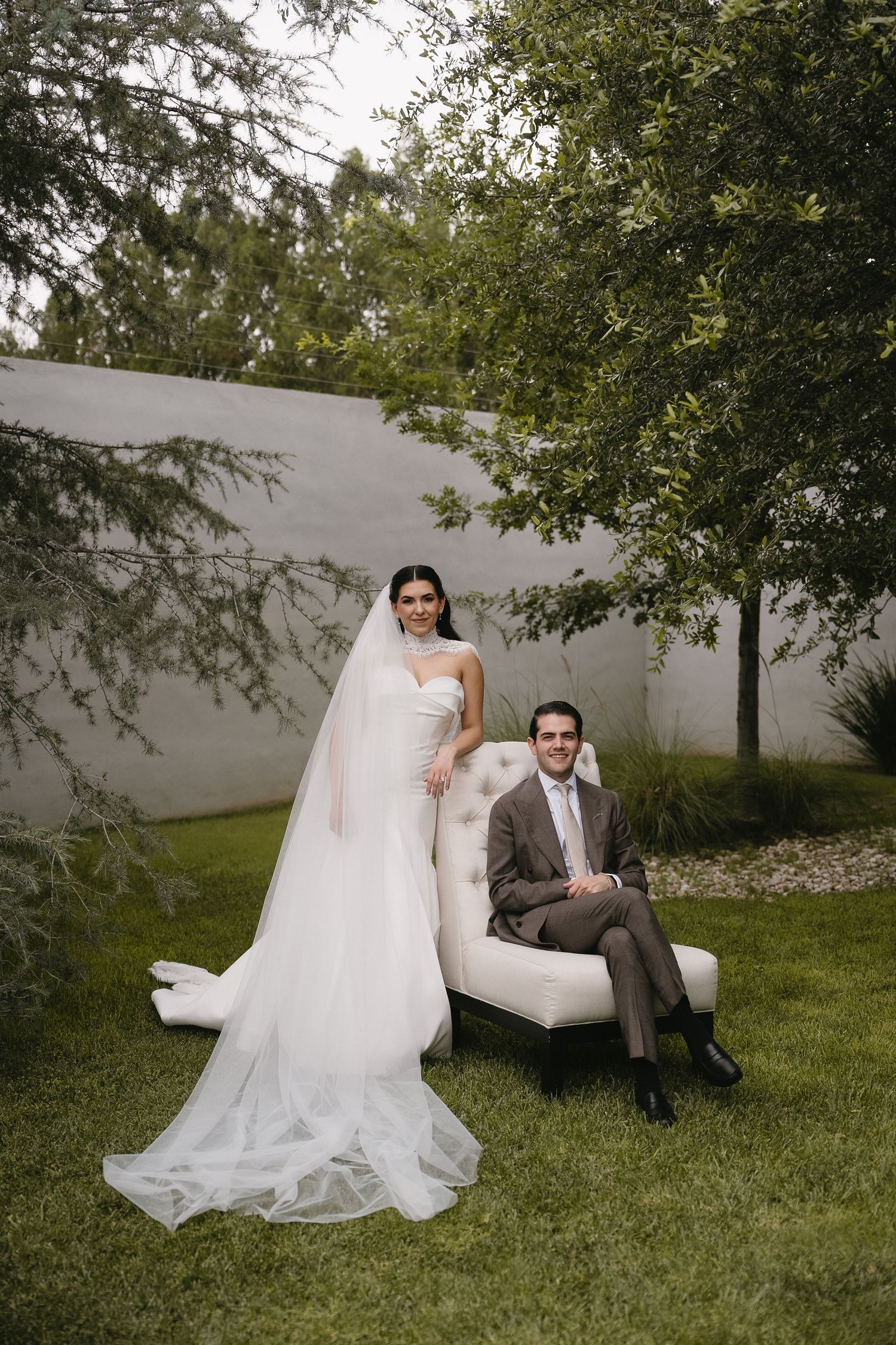 Bride in white gown with veil stands beside groom in tan suit seated on tufted chair in lush garden.