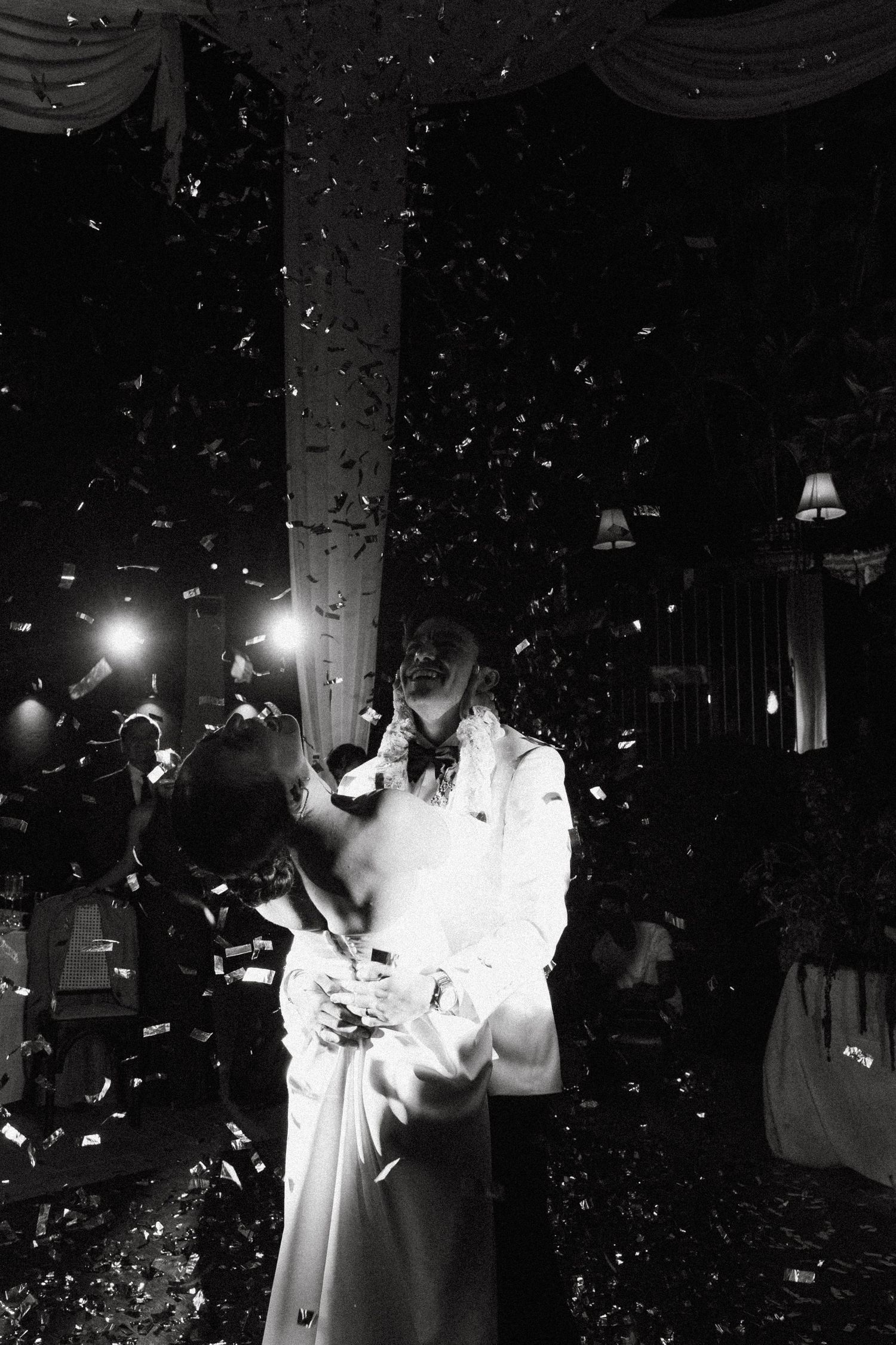 Black and white photo of a wedding celebration with confetti falling in a dimly lit indoor venue.