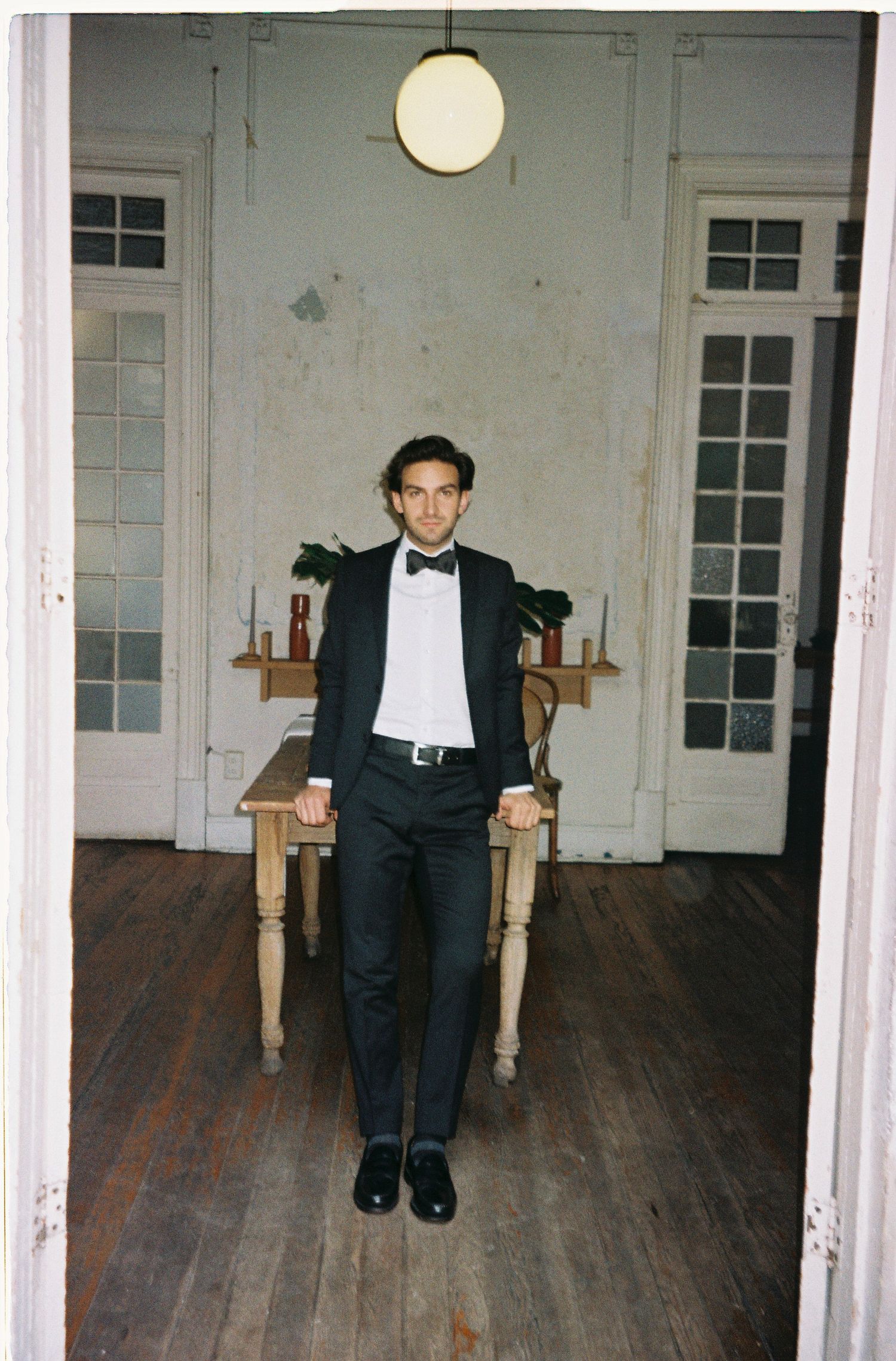 Groom in classic black tuxedo poses casually in vintage room setting.