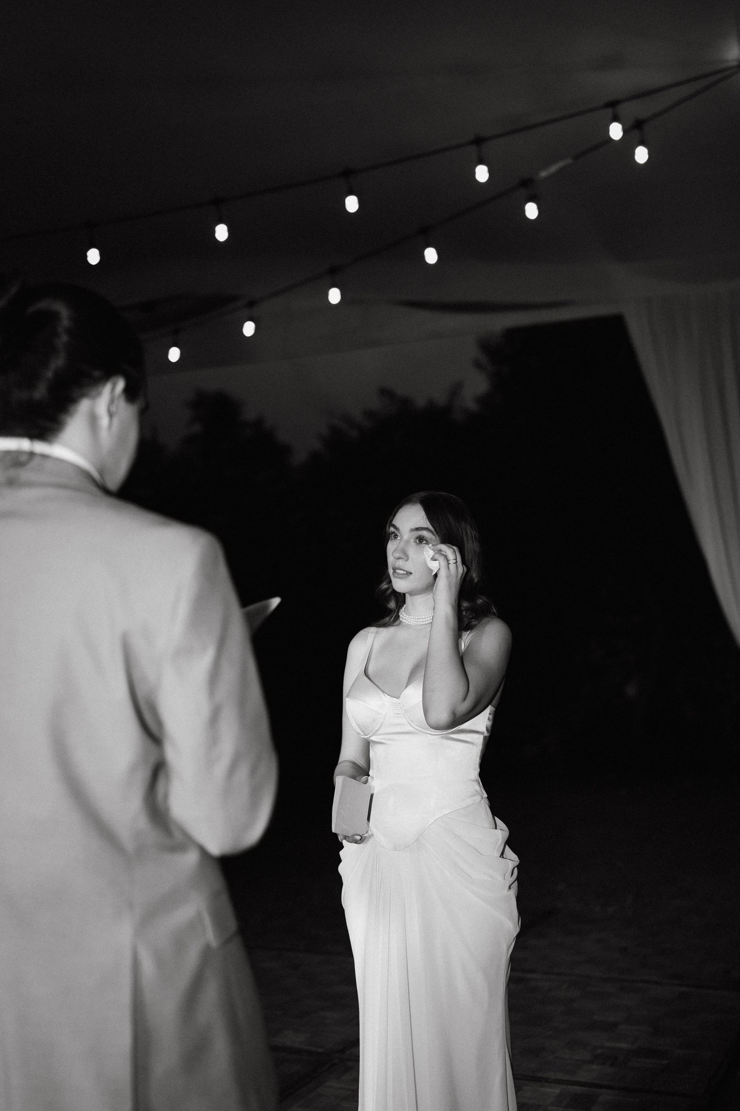 Outdoor nighttime scene with string lights and white-clad figures in conversation.