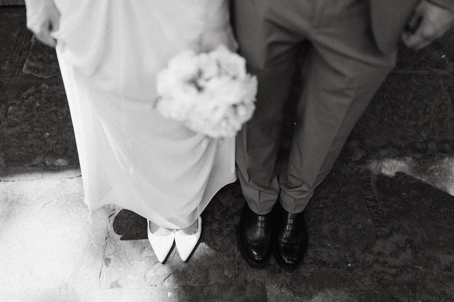 Black and white photo showing the bottom of a wedding dress and formal shoes.