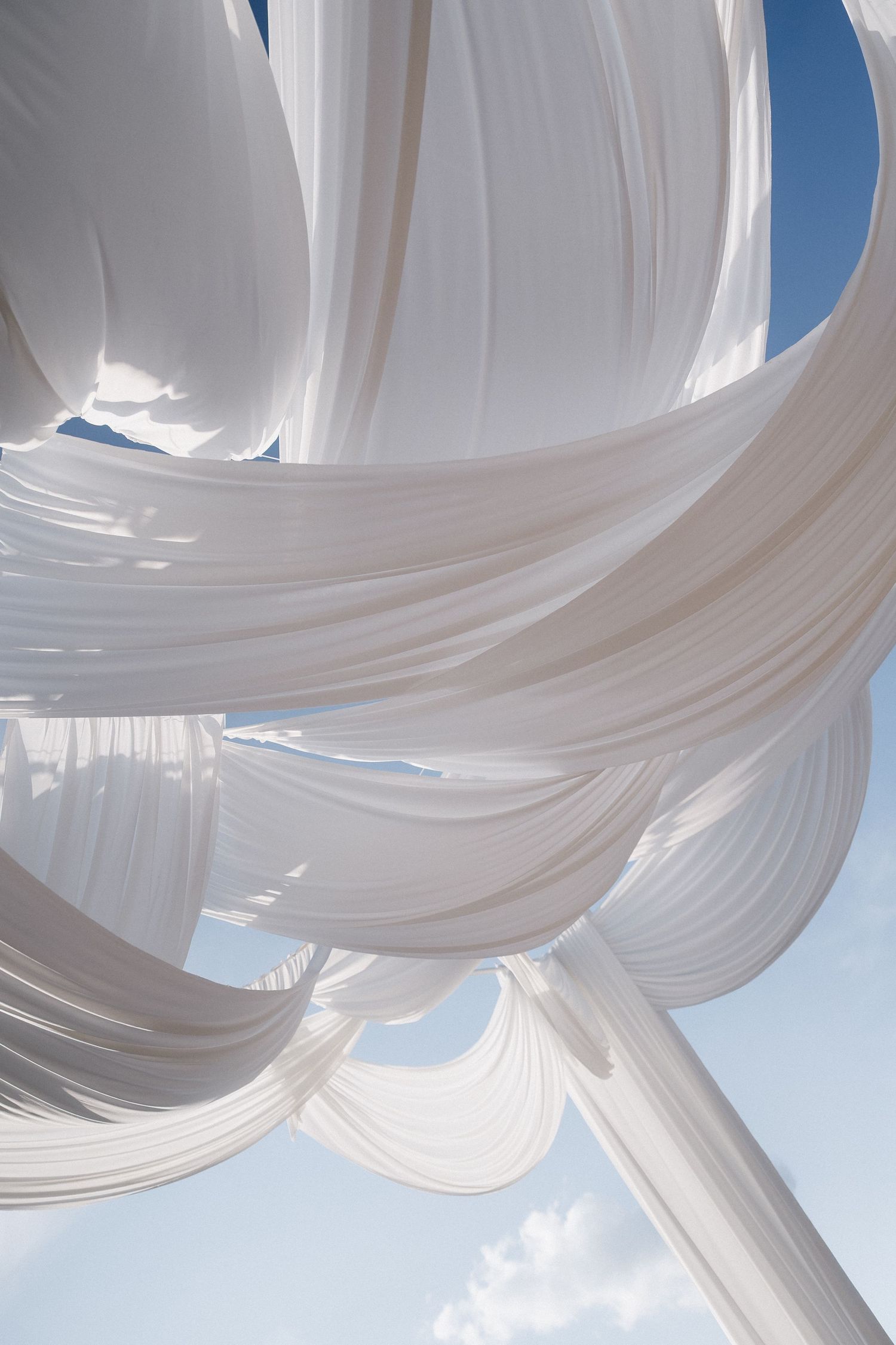 Flowing white fabric drapes billow elegantly against a bright blue sky with wispy clouds.