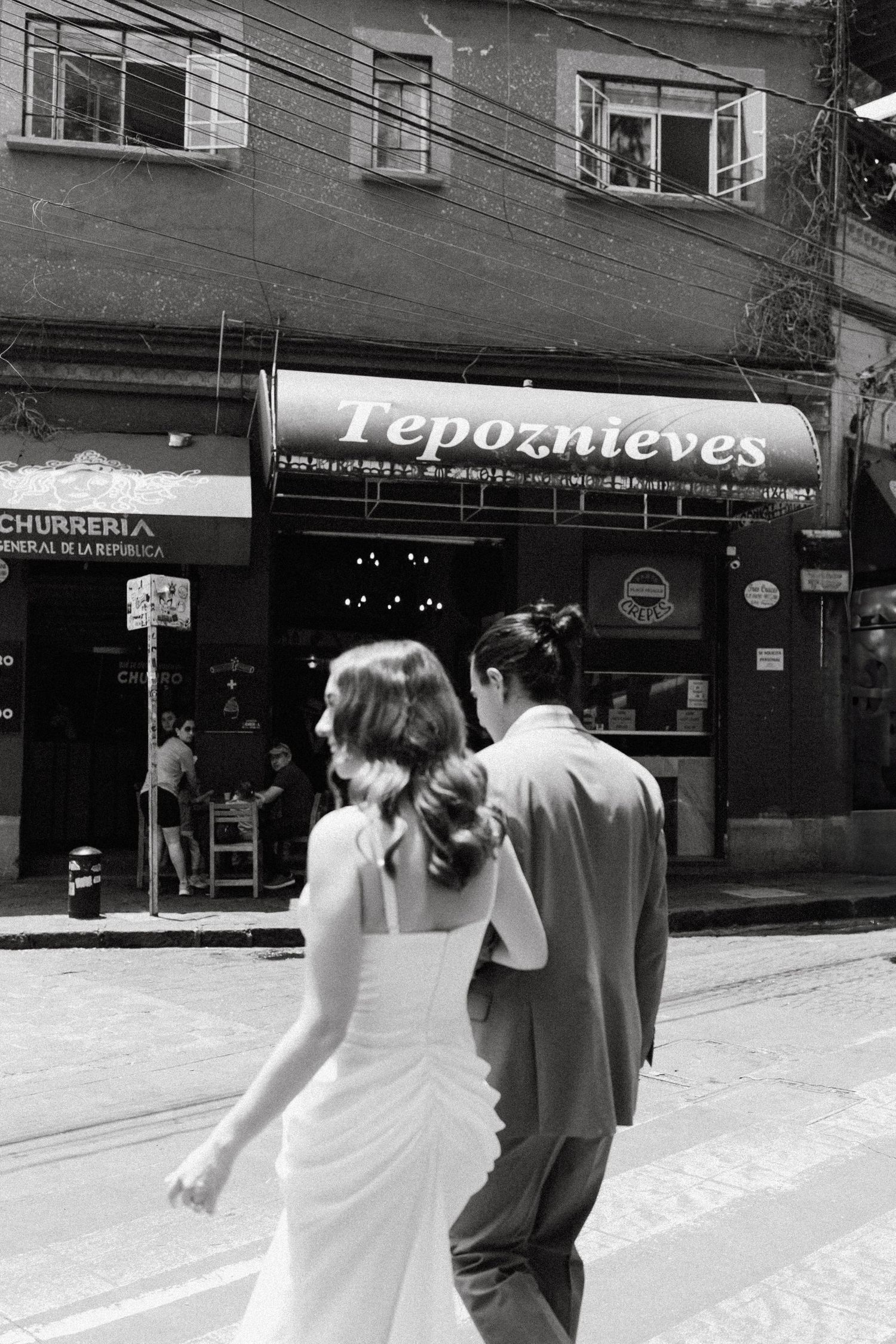 A couple strolls down a street past shops in black and white photography.
