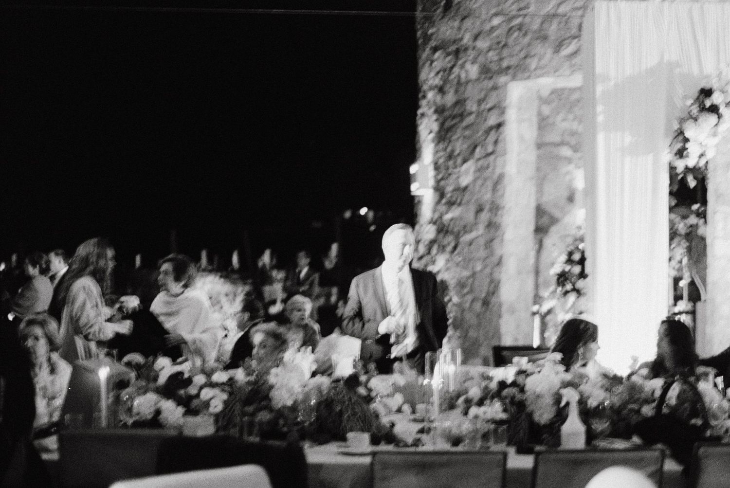 A black and white photograph shows people gathered at an elegant dinner event with floral centerpieces and formal decor.
