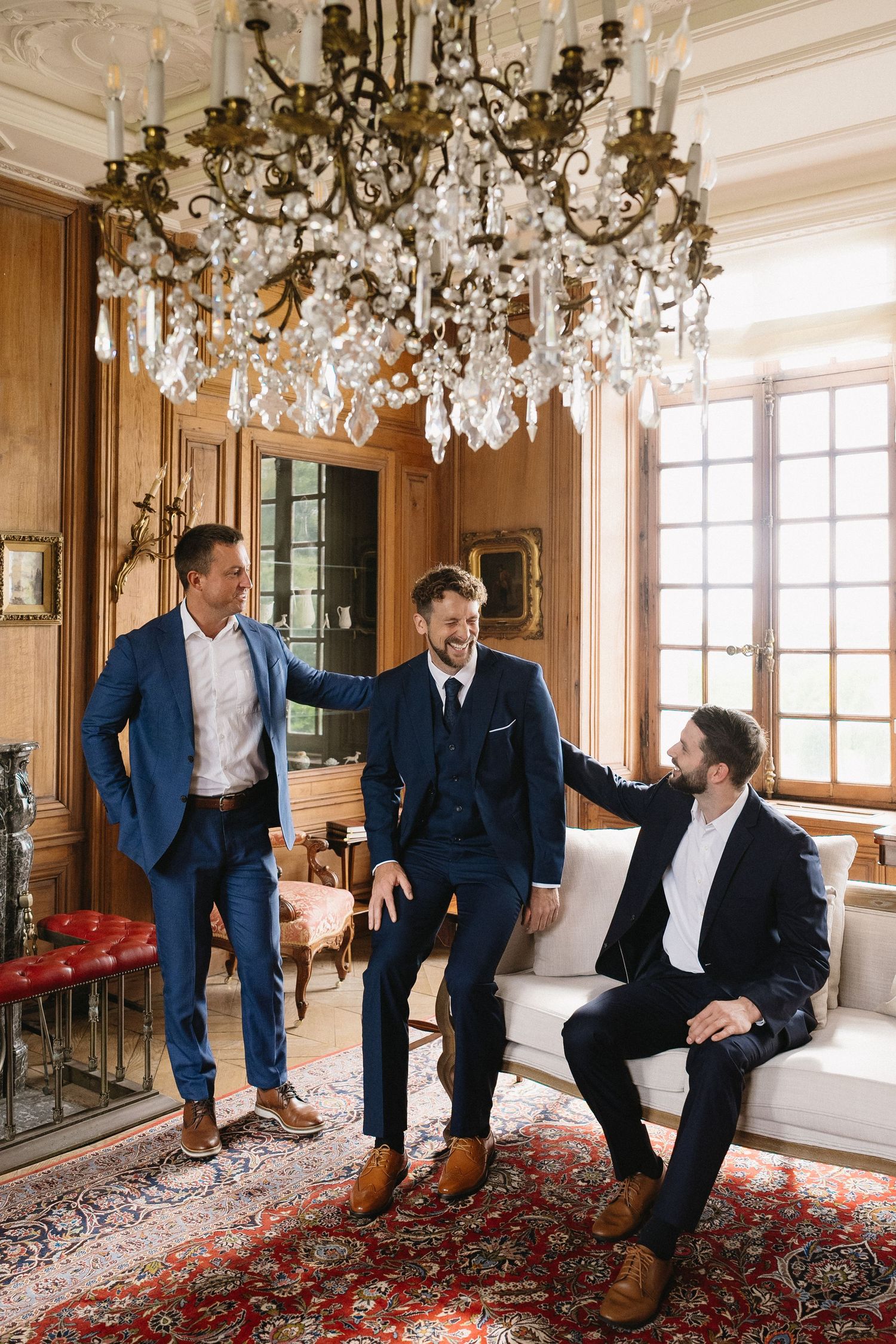 Three groomsmen in navy suits relaxing under an ornate crystal chandelier in a luxurious room.