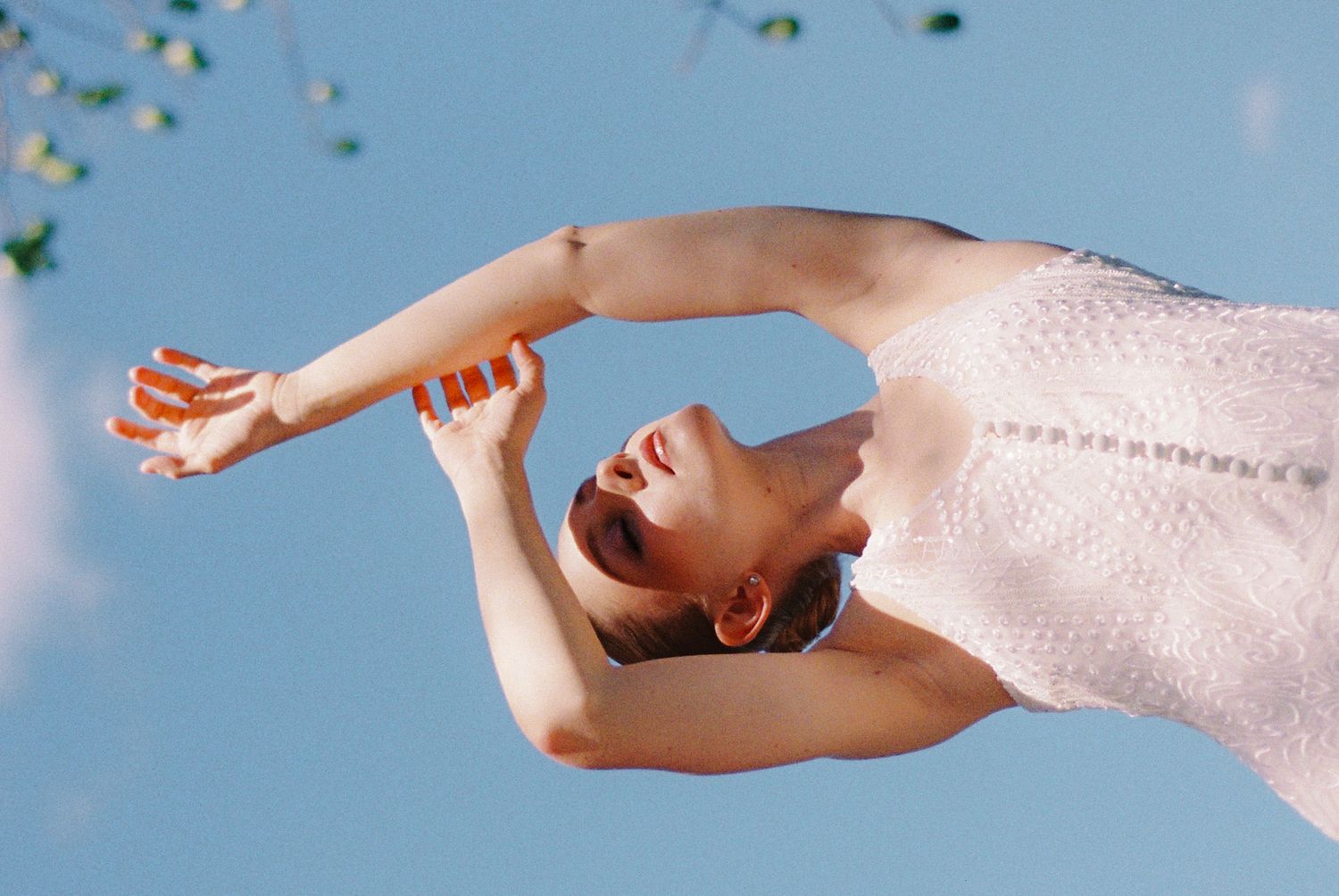 A woman in a pink dress gracefully arches backward against a clear blue sky, arms extended elegantly.