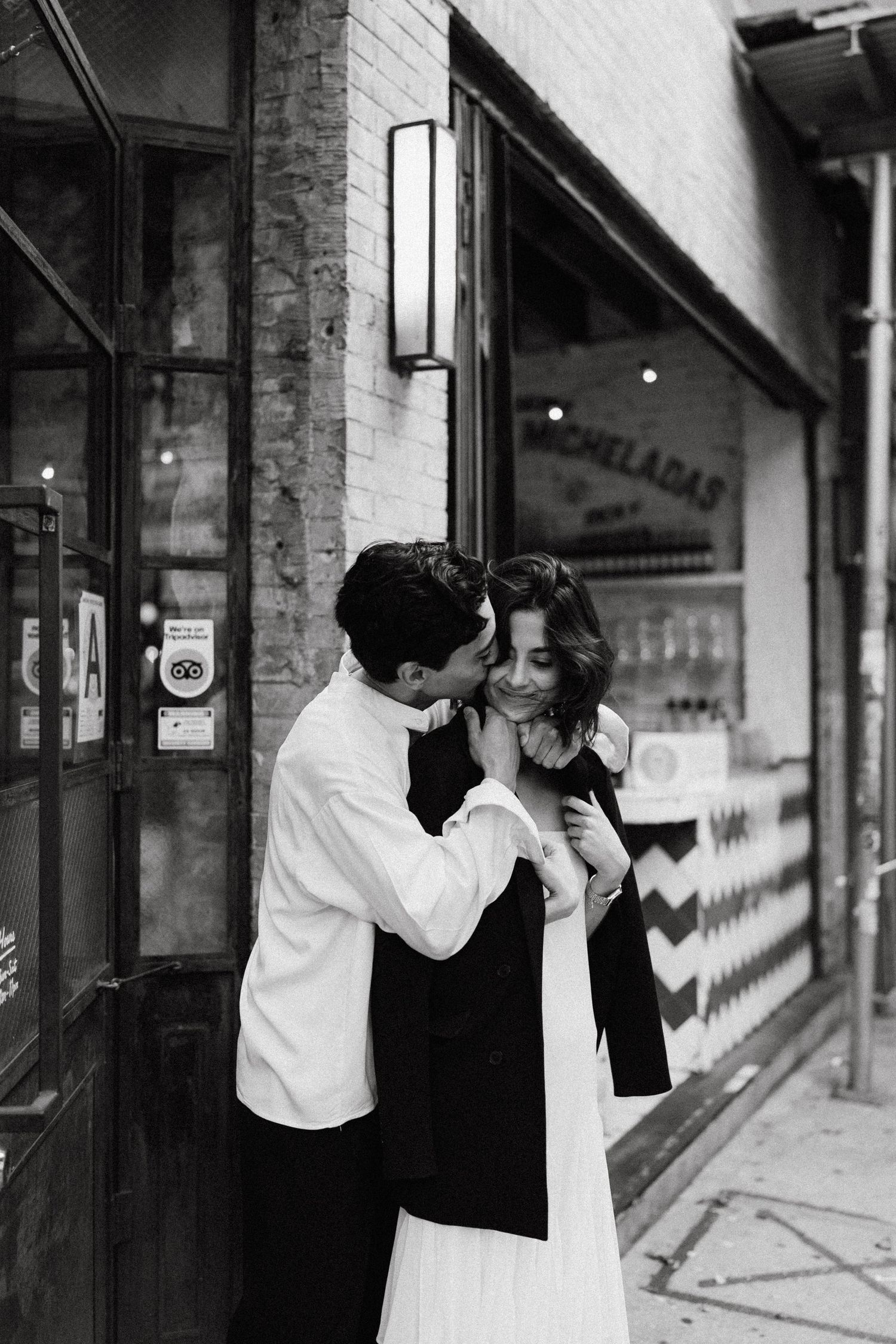 A couple shares a tender moment in black and white against a brick storefront.