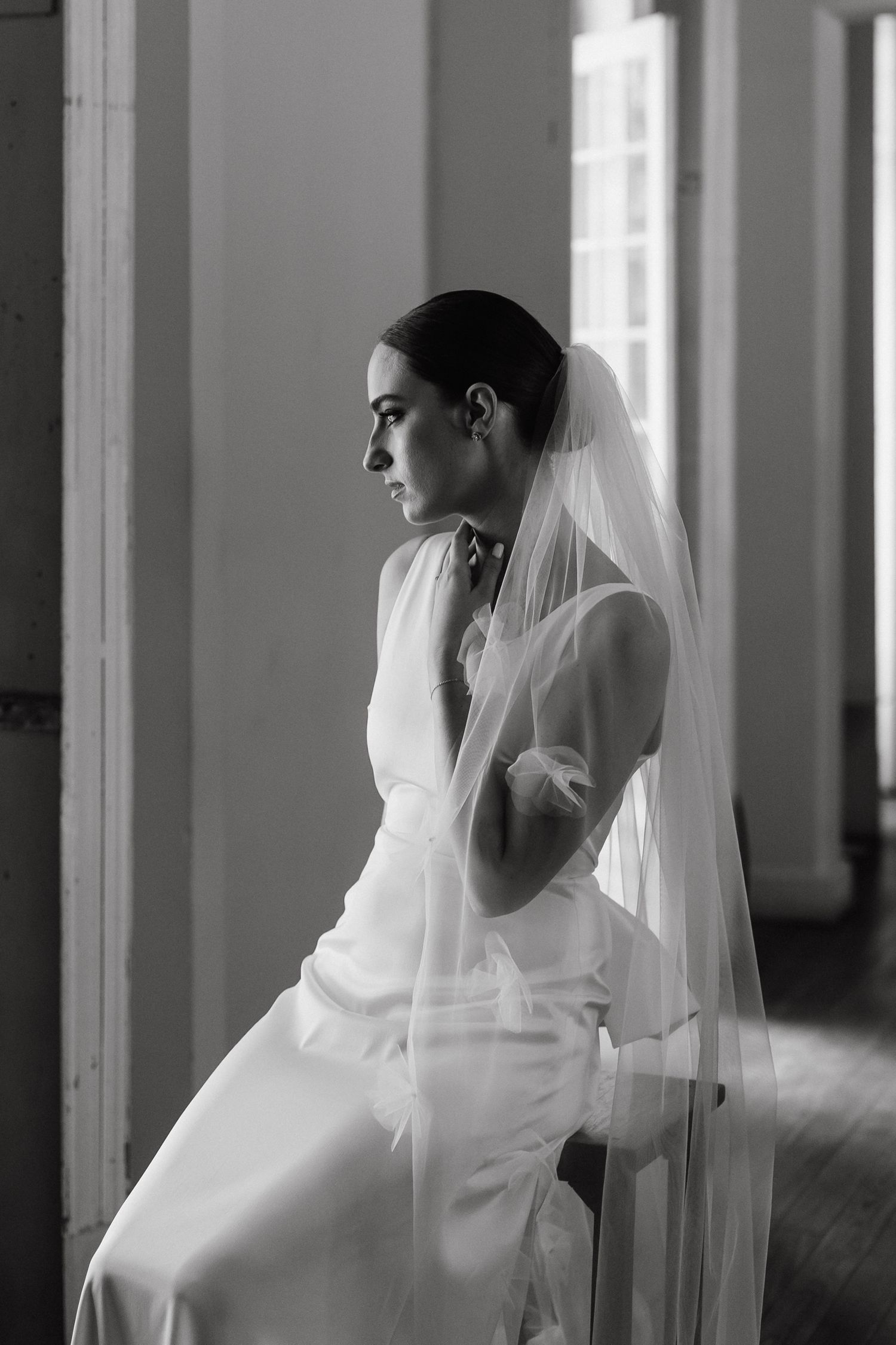 Black and white portrait of bride in sleek dress and veil gazing towards window.