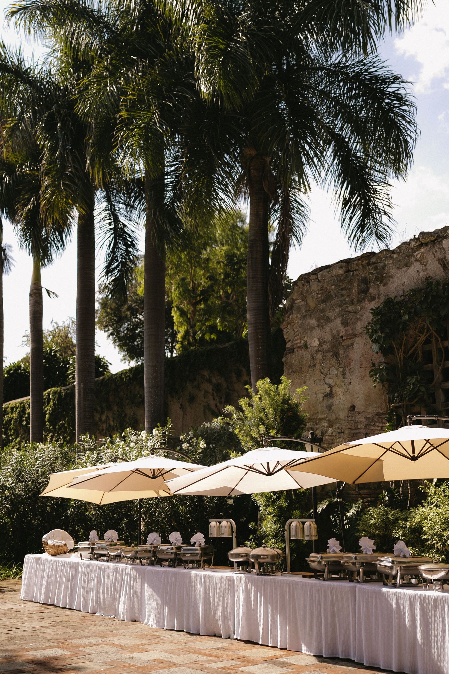 Elegant outdoor buffet setup with white tablecloths and beige umbrellas against stone walls and tall palm trees.