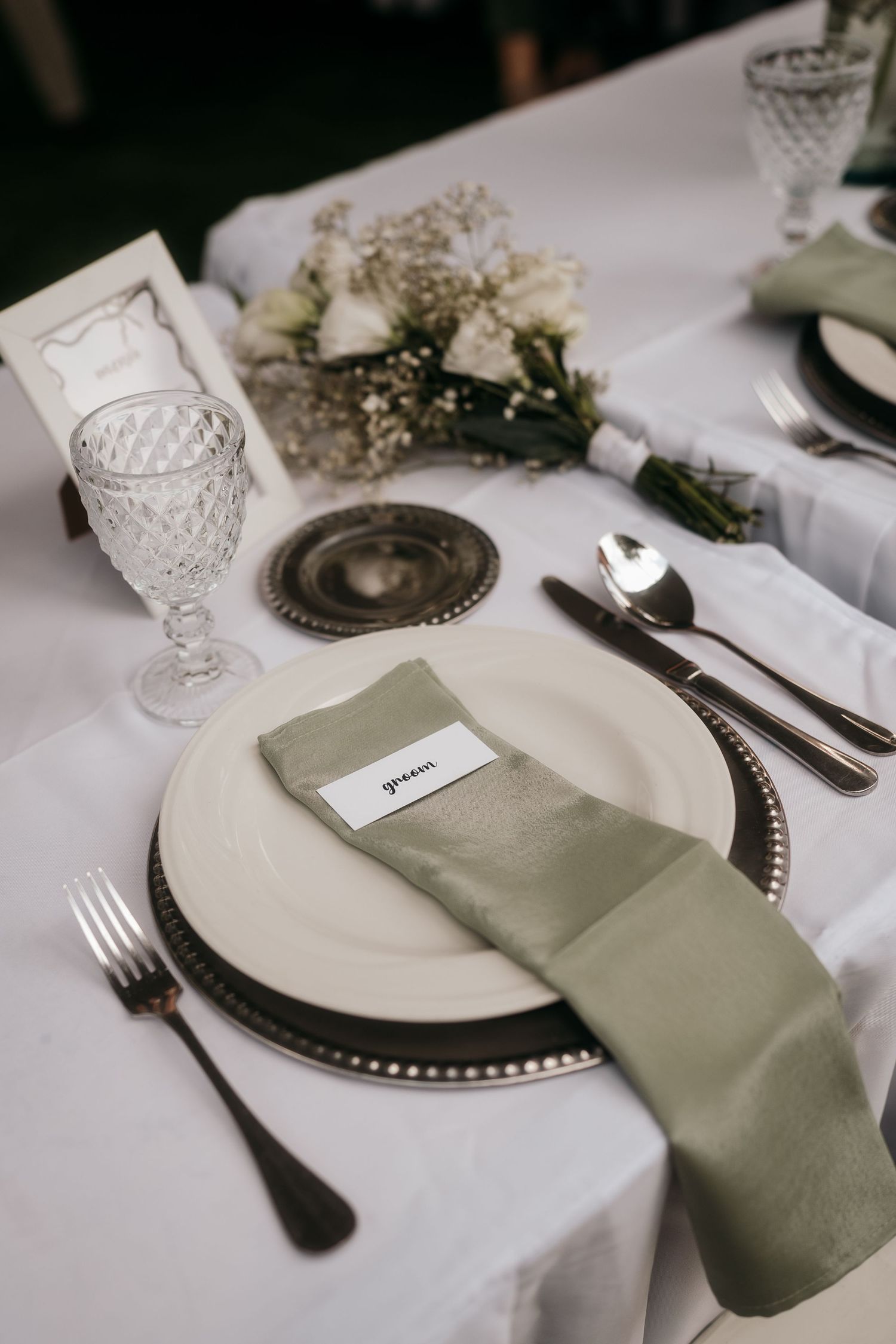 Elegant place setting with white plate, sage napkin, and crystal glassware on white tablecloth.