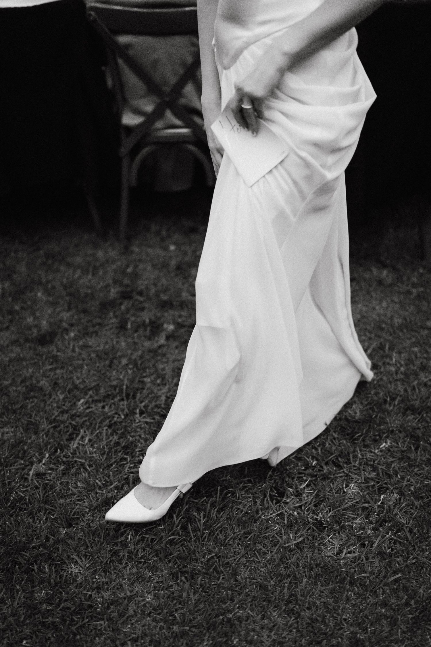 Black and white detail shot of flowing white wedding dress fabric against grass.