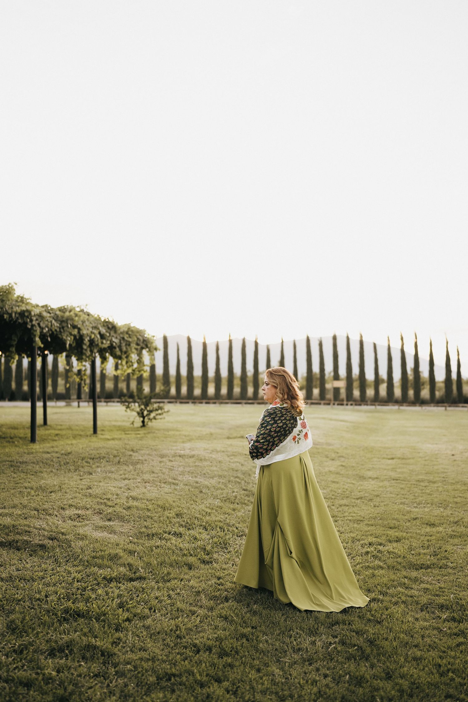 A person in a lime green dress stands in a grassy field with cypress trees lining the horizon.