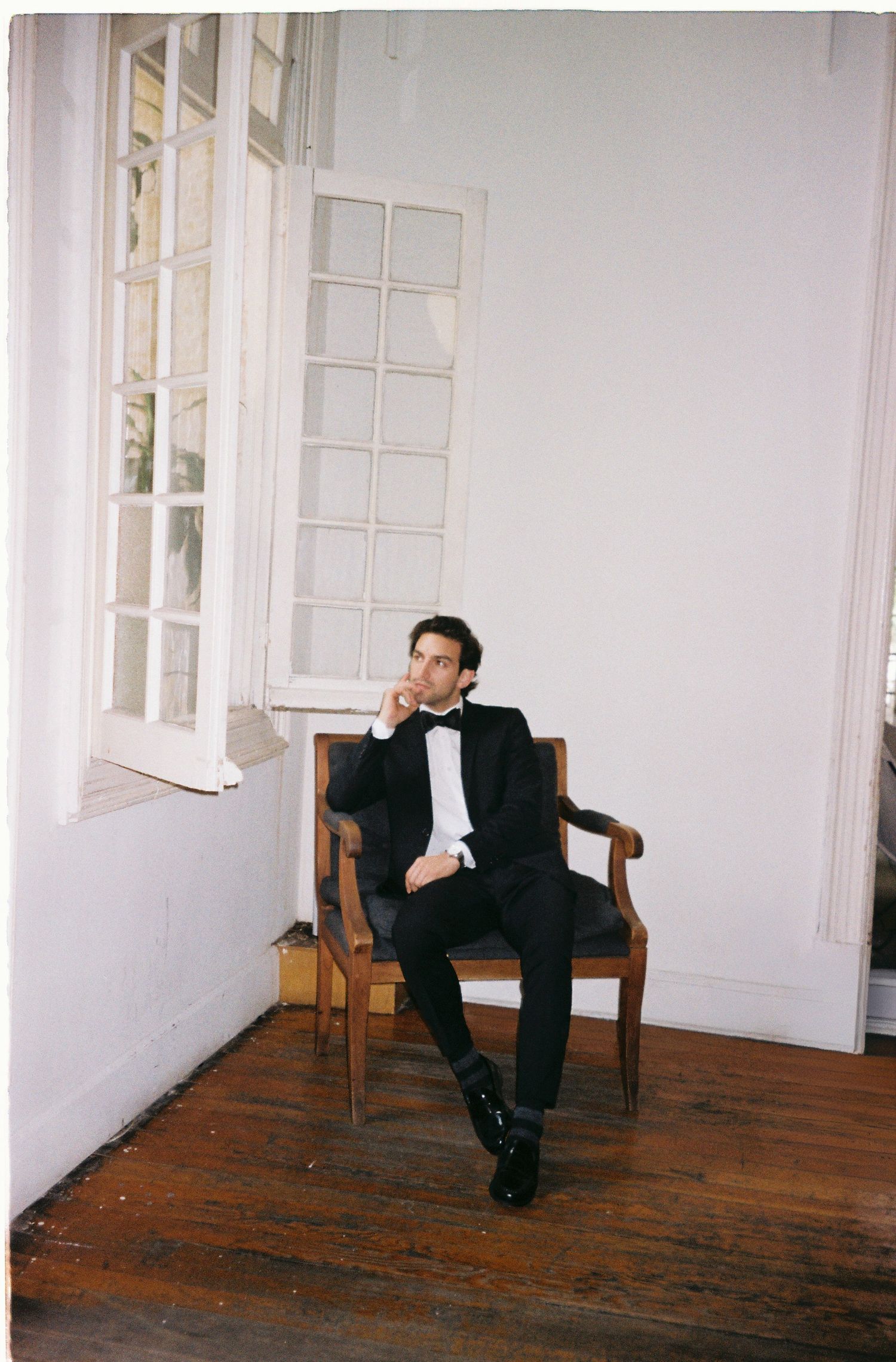 Candid portrait of groom relaxing in wooden chair by window.