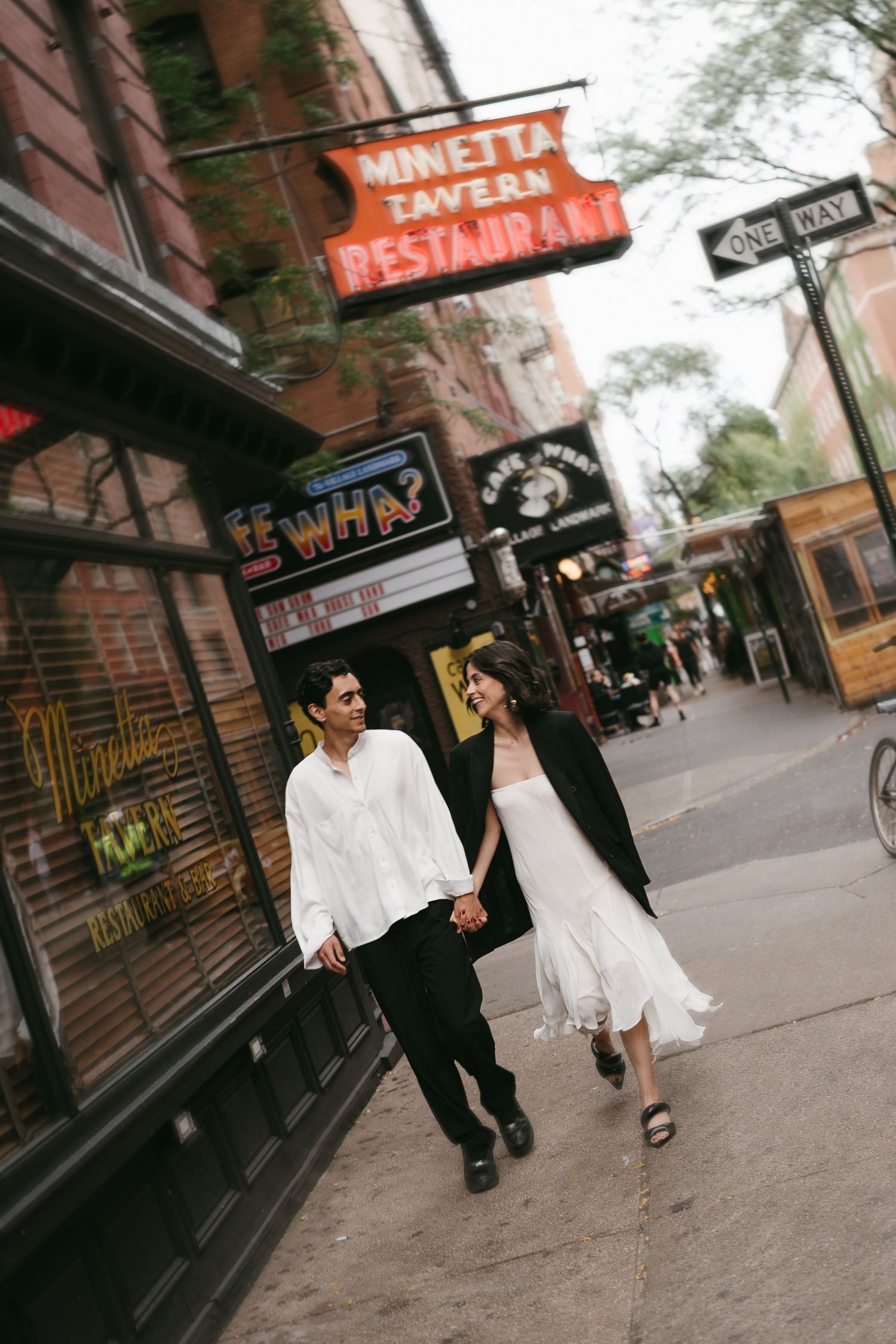 Couple walking past storefronts on urban street in casual elegant attire.