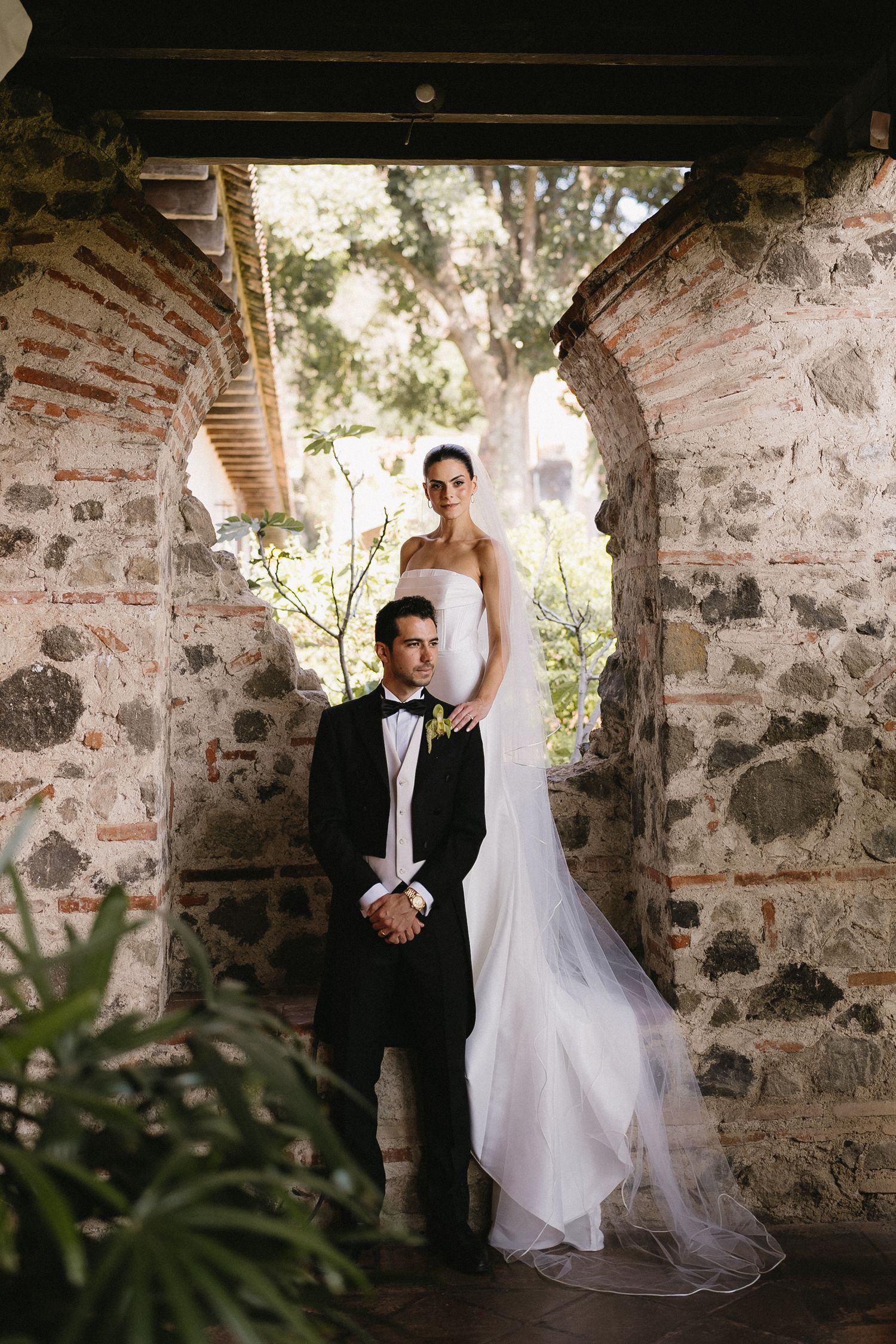 Bride in strapless gown and groom in black tuxedo pose elegantly in a rustic stone archway surrounded by lush greenery.