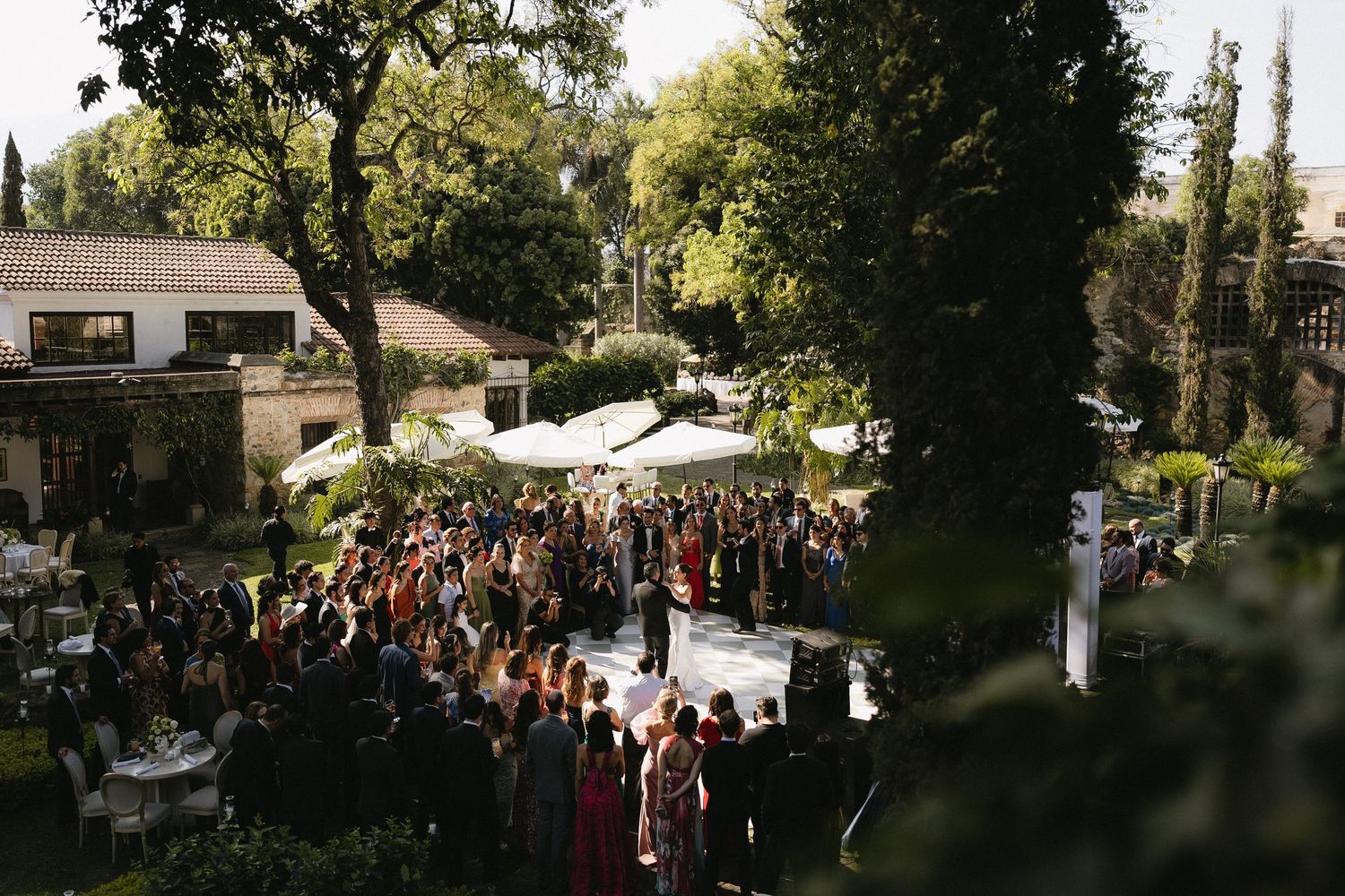 Aerial view of an outdoor wedding ceremony with guests gathered around a white platform under trees.