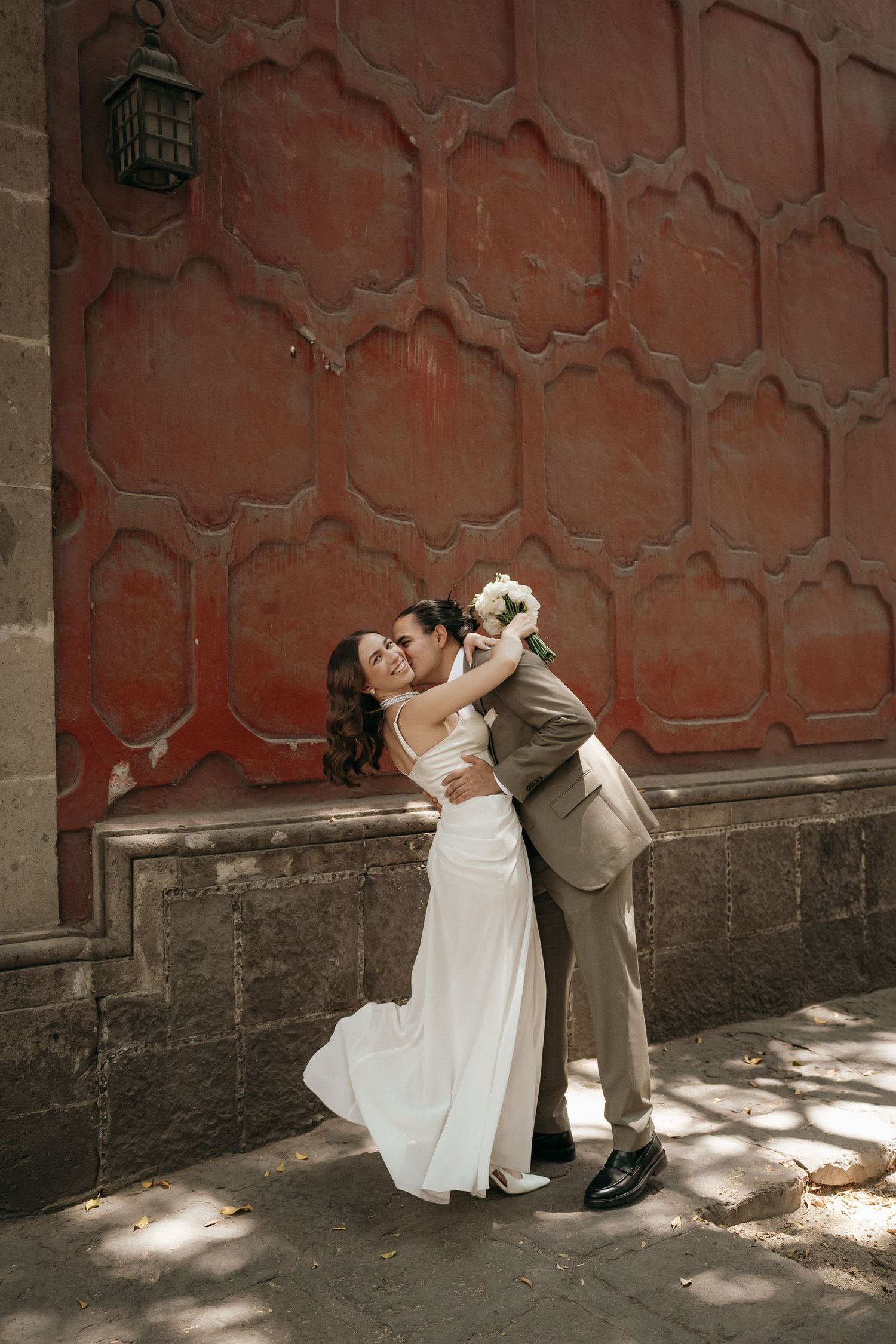 A couple poses dramatically against a textured red wall in wedding attire.