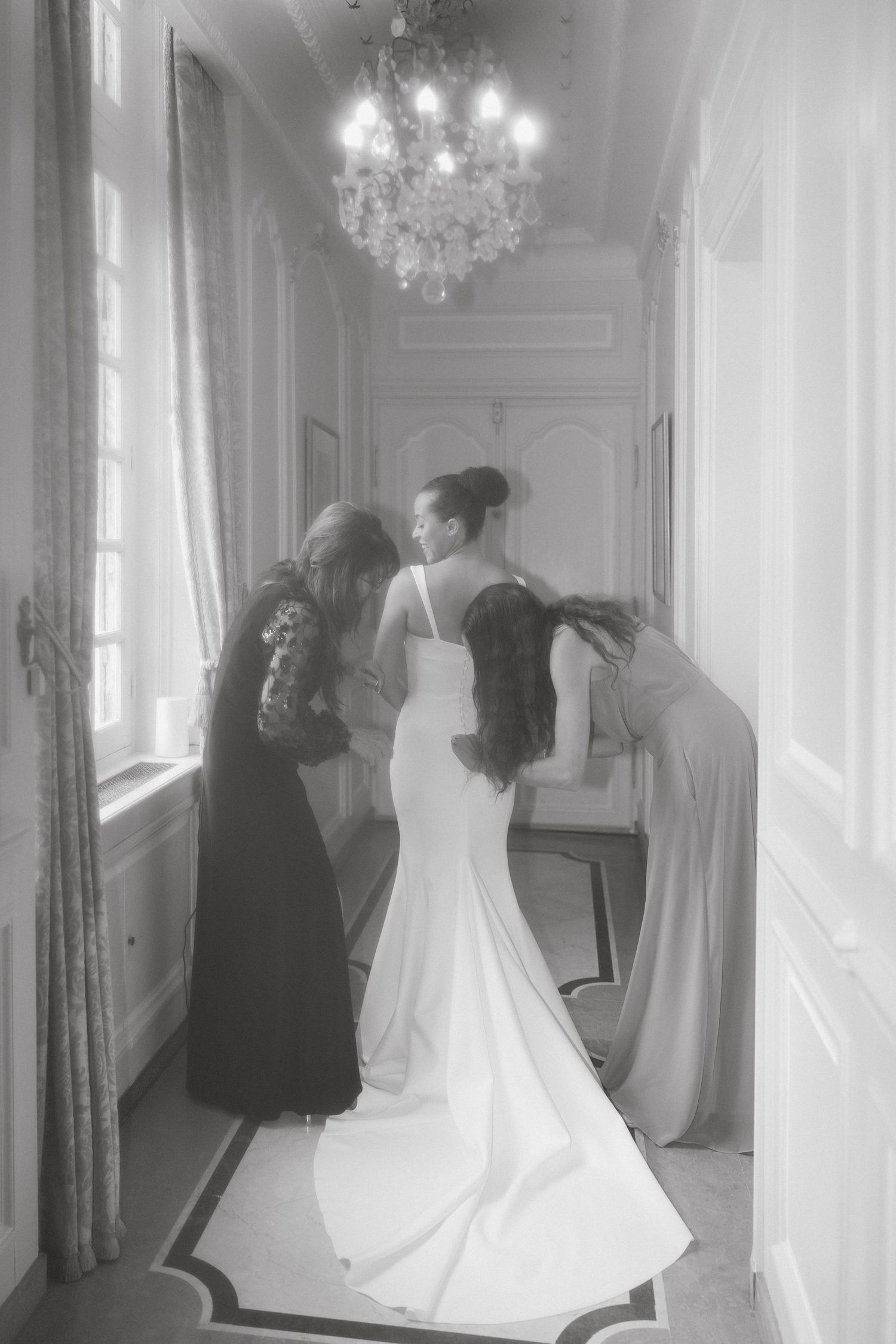 A bride in a white mermaid gown being helped with her dress in an elegant hallway with a crystal chandelier.