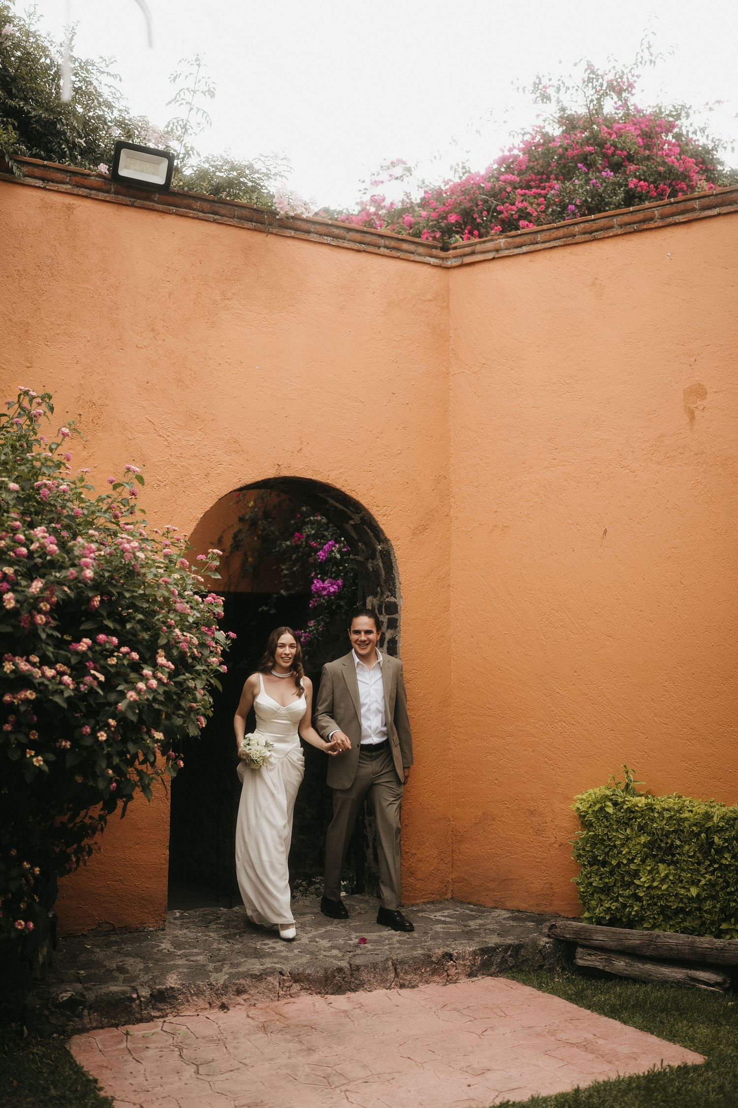 A couple emerges through an arched doorway in a peach-colored courtyard with blooming flowers.