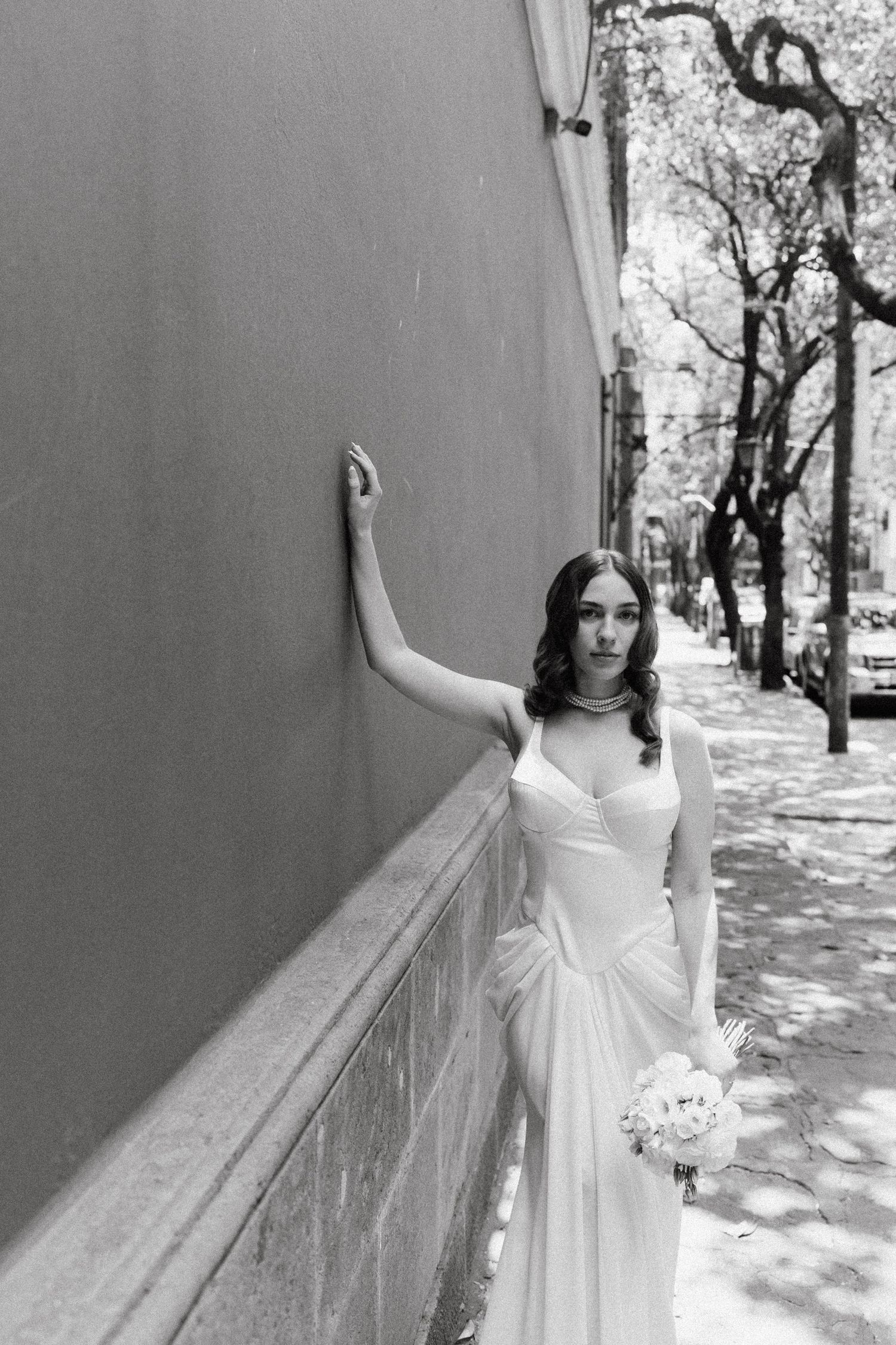 A bride in white poses elegantly against a gray wall in black and white photography.