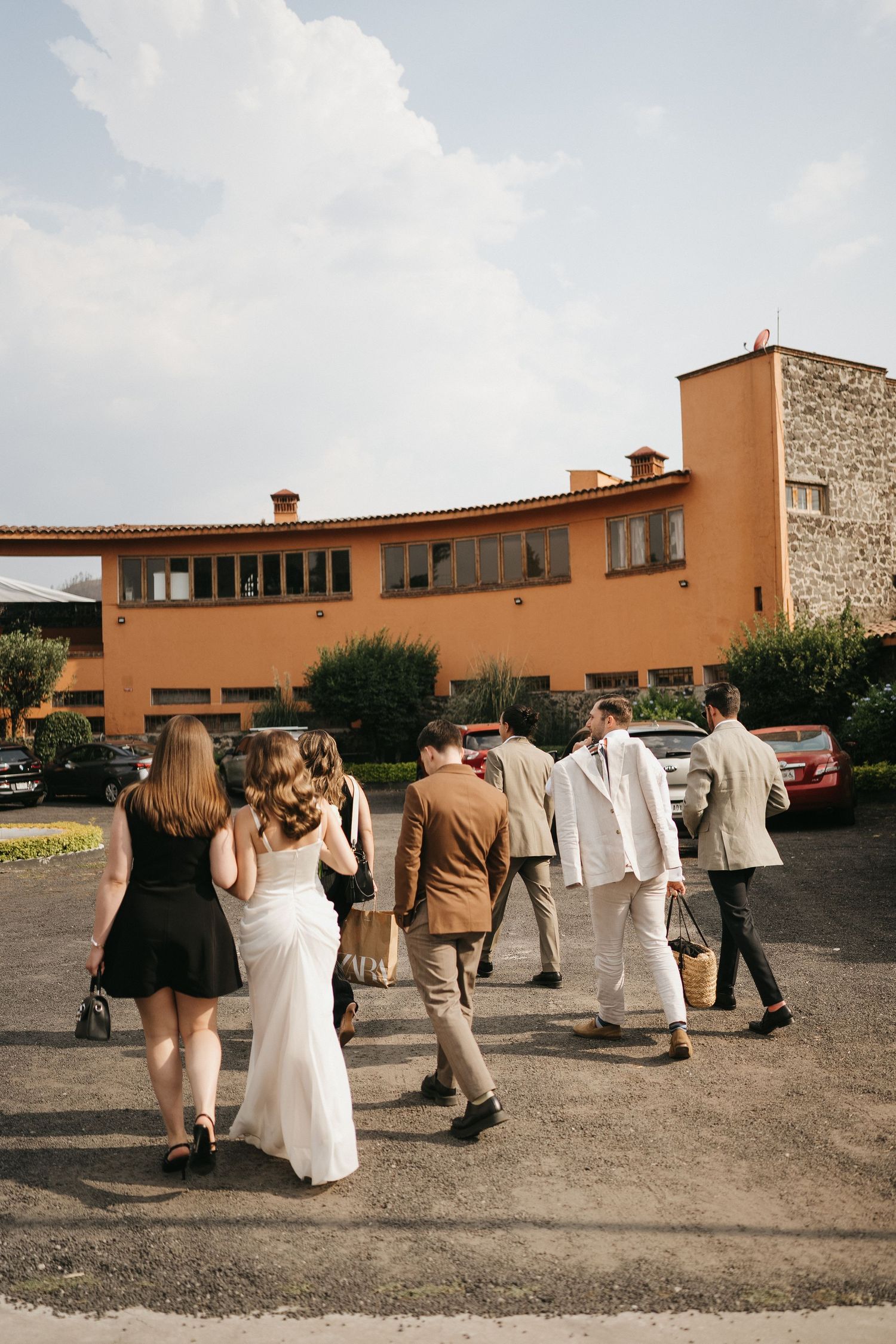 Wedding guests in formal attire walk in a line outside a terracotta-colored building.