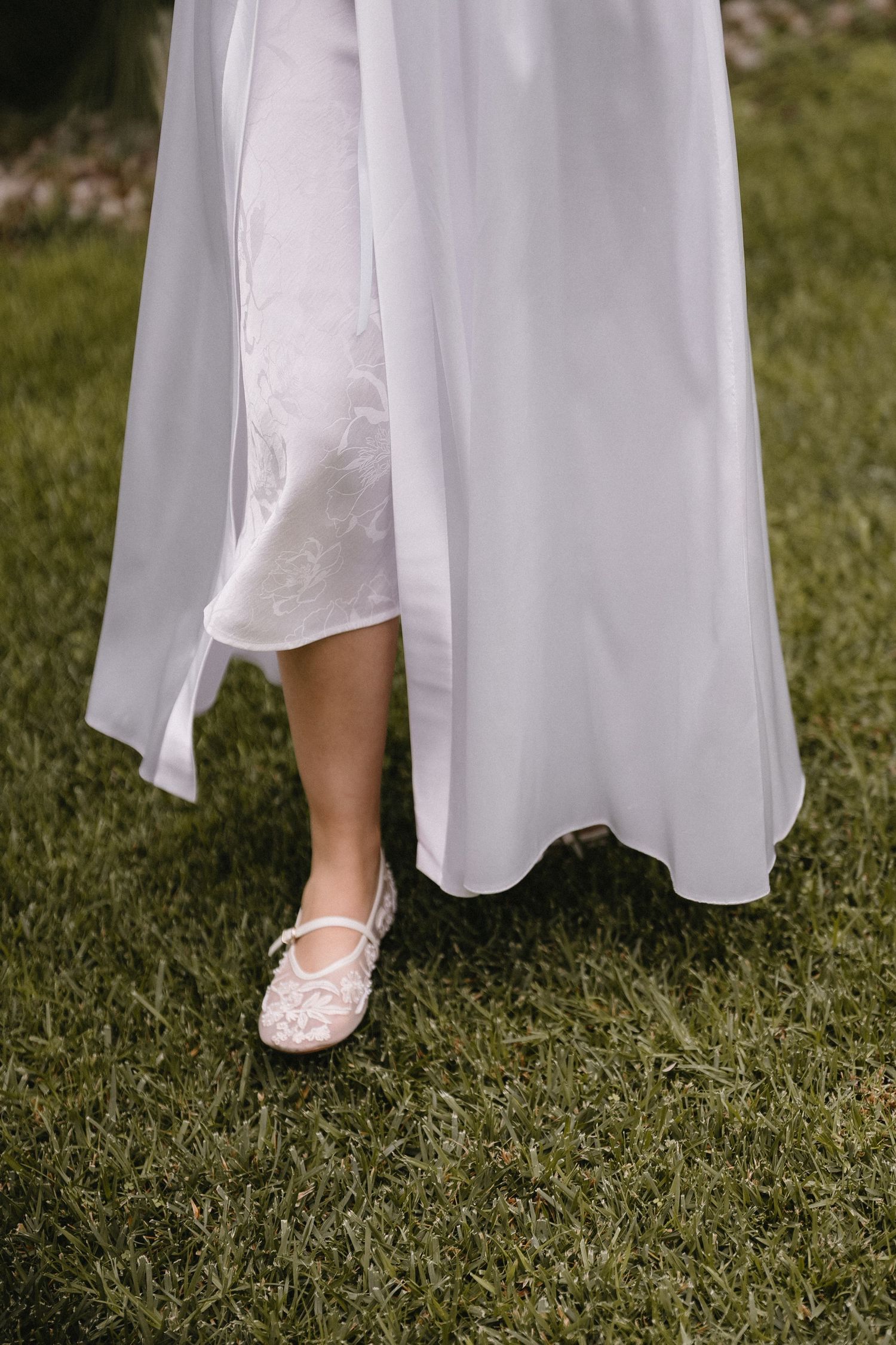 White bridal shoes peek out beneath a flowing dress on green grass.