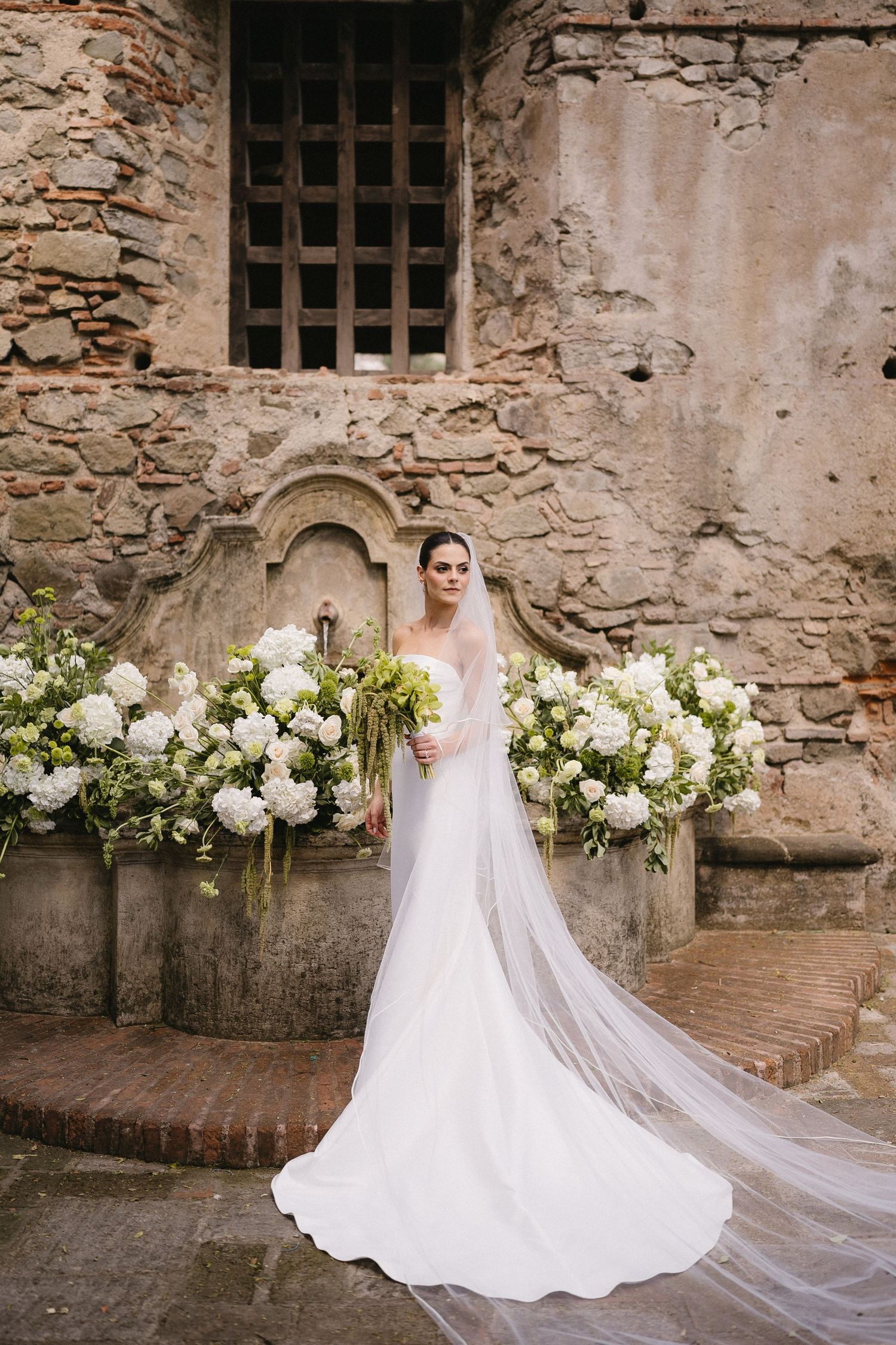 Elegant white wedding dress with flowing veil against rustic stone wall adorned with white floral arrangements.