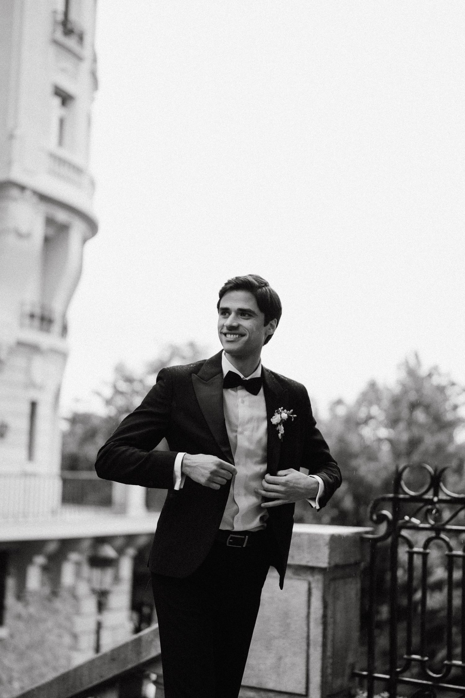 Person in formal attire standing on a balcony with ornate railings in black and white.