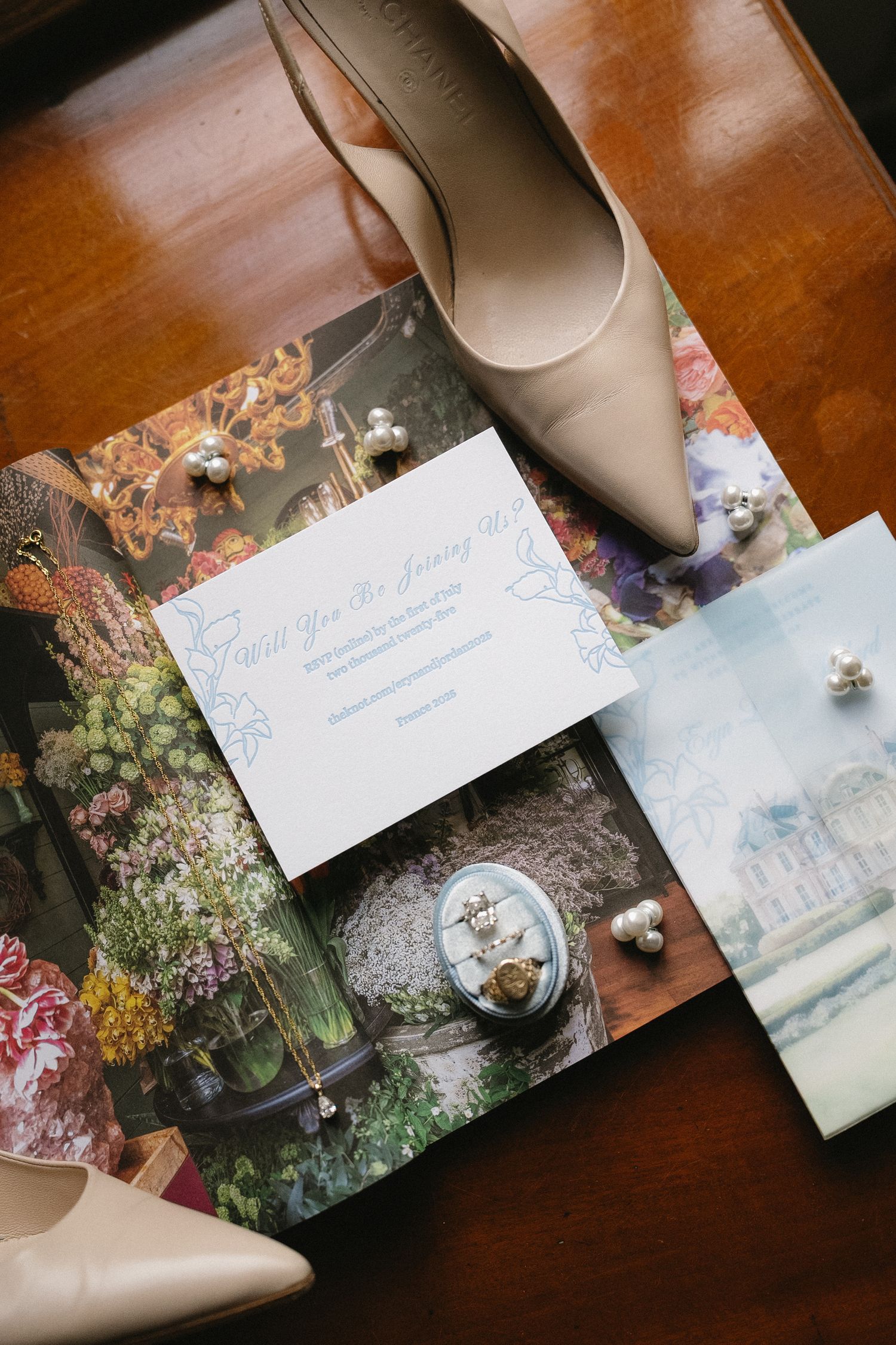 Wedding stationery and nude shoes arranged artistically on wooden surface with floral accents.