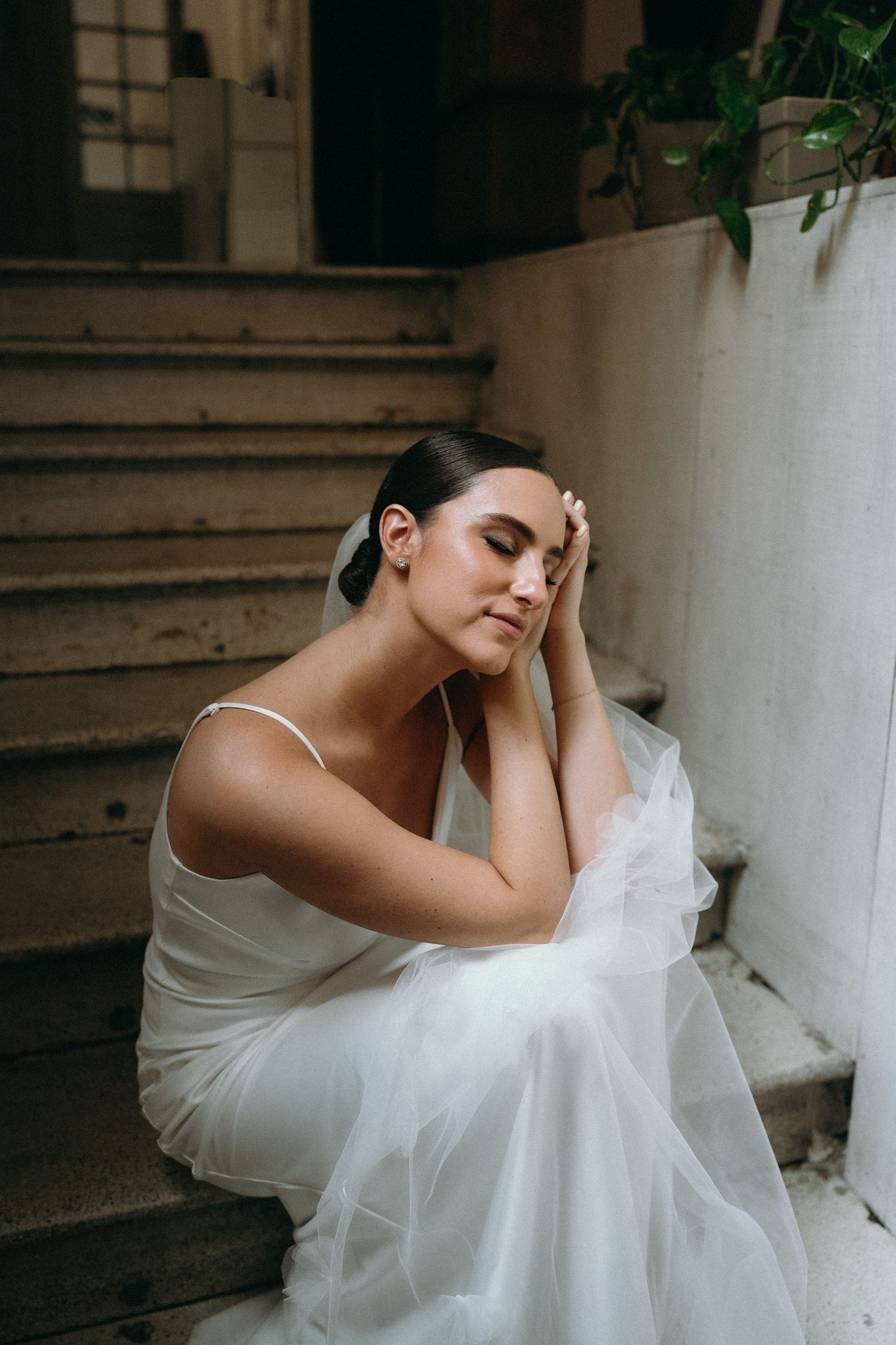 A bride in a white dress sits elegantly on concrete stairs in a romantic pose.