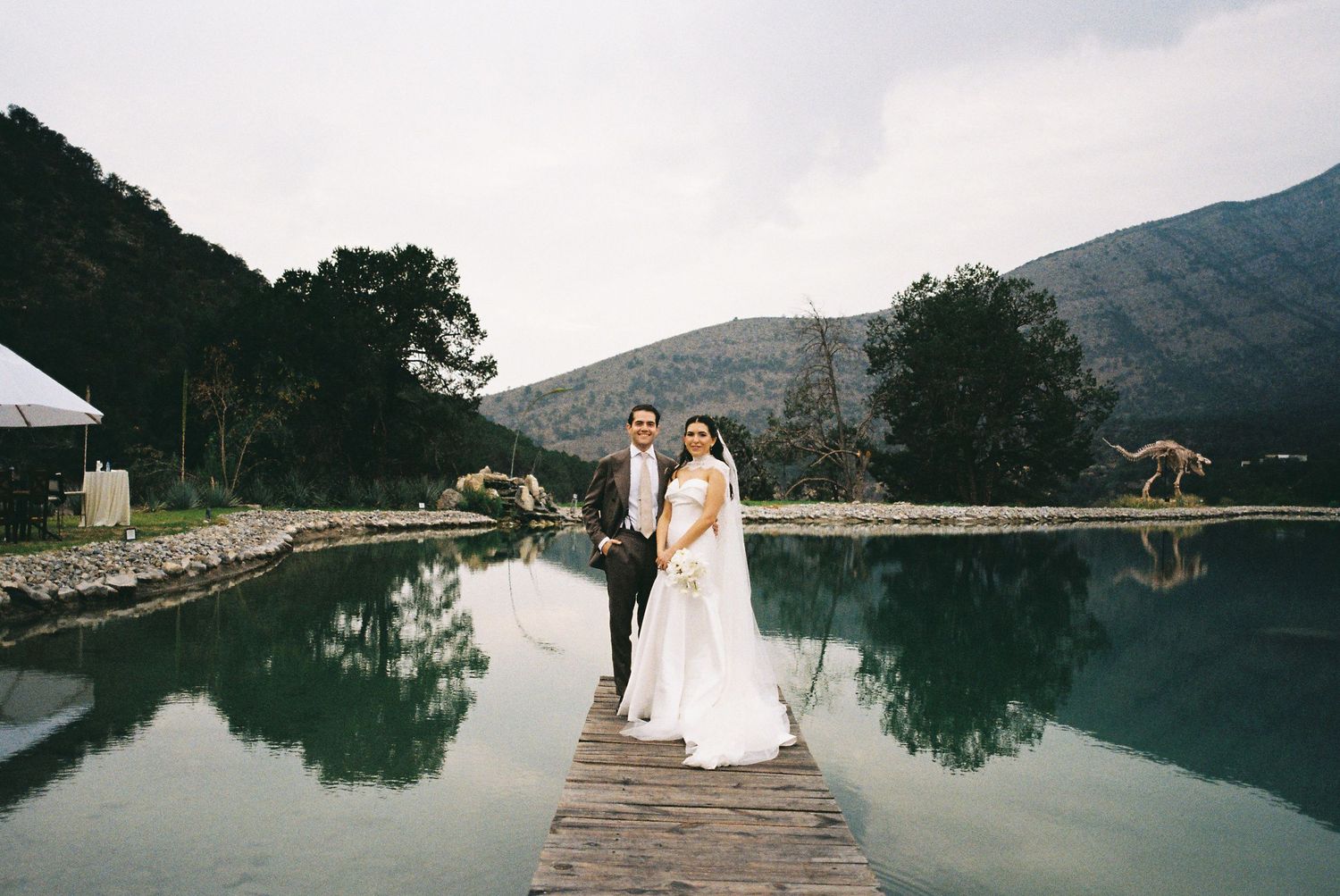 Wedding couple poses on wooden dock overlooking serene mountain lake at sunset.