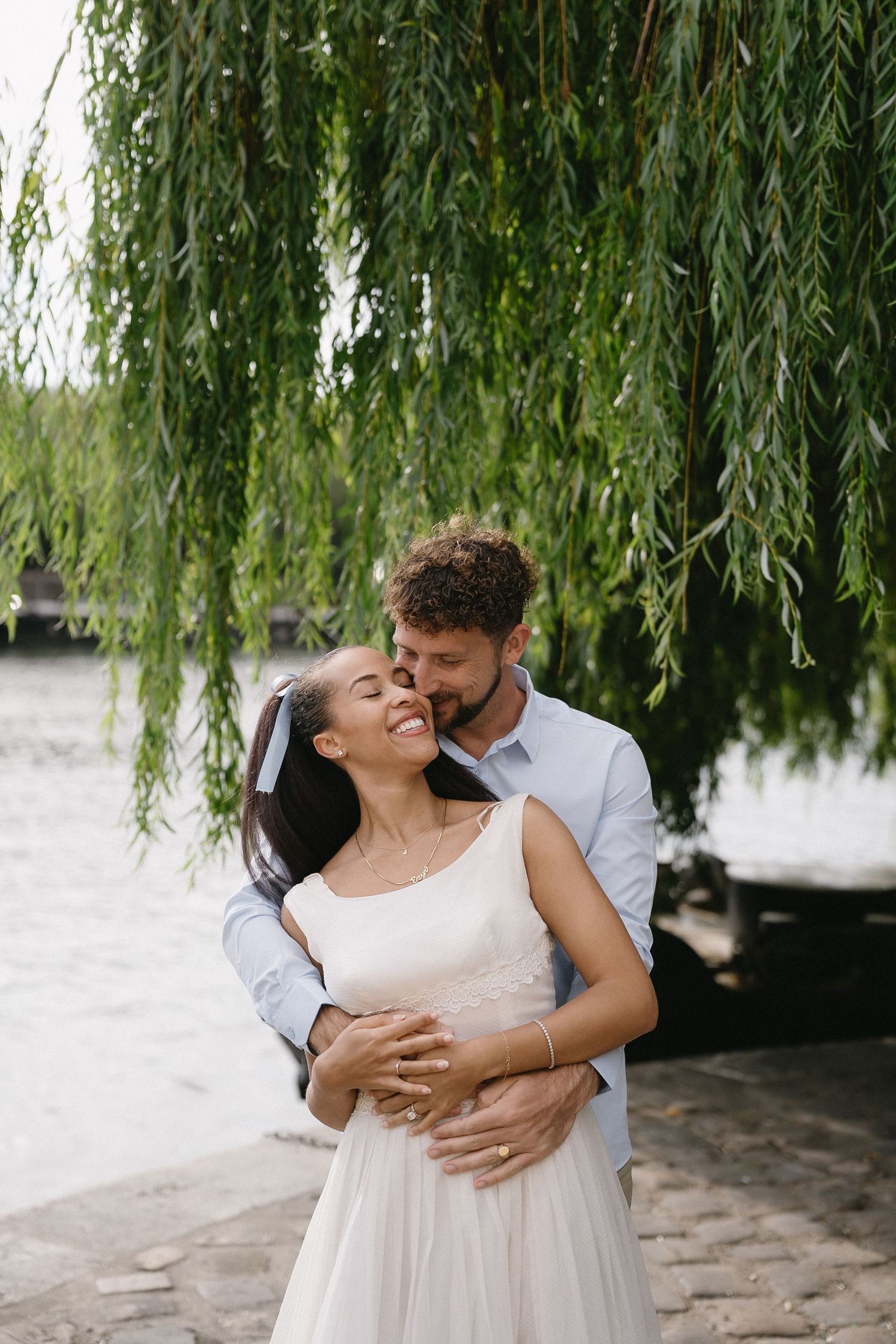 Couple embracing under a weeping willow tree by the water.