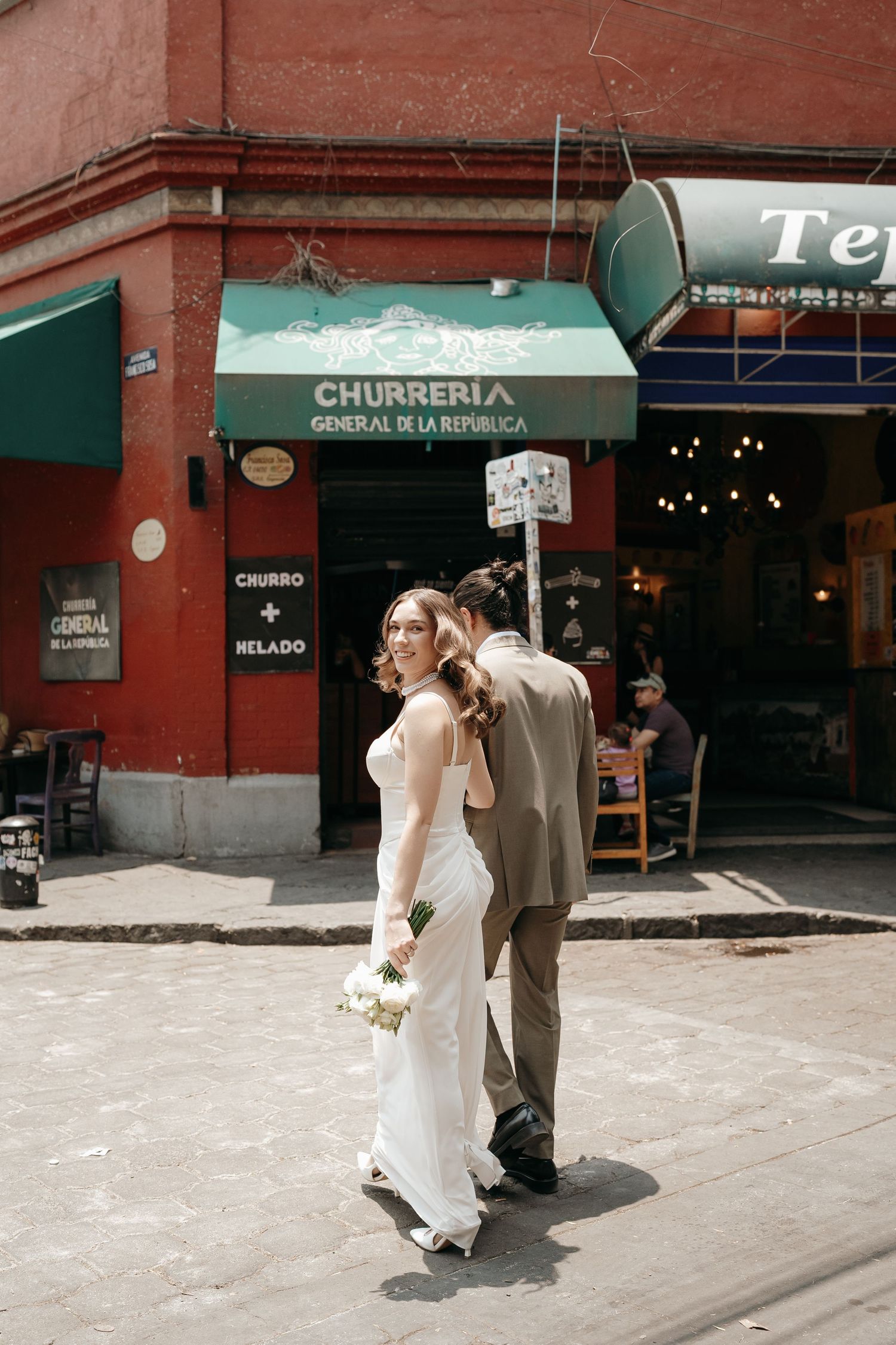 A couple walks together past a churreria storefront on a sunny street.