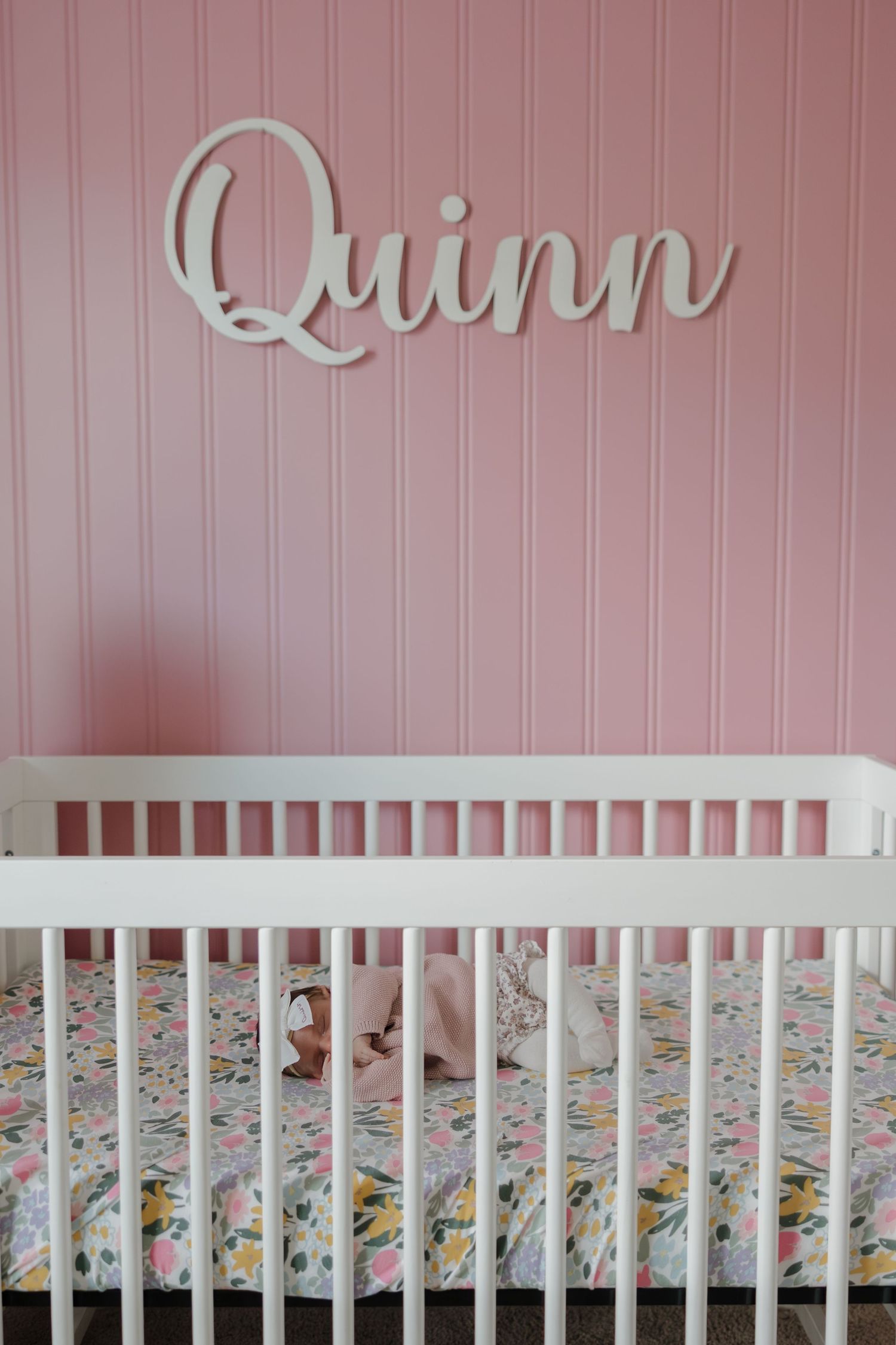 White crib with floral bedding set against a pink wall with decorative name lettering mounted above.