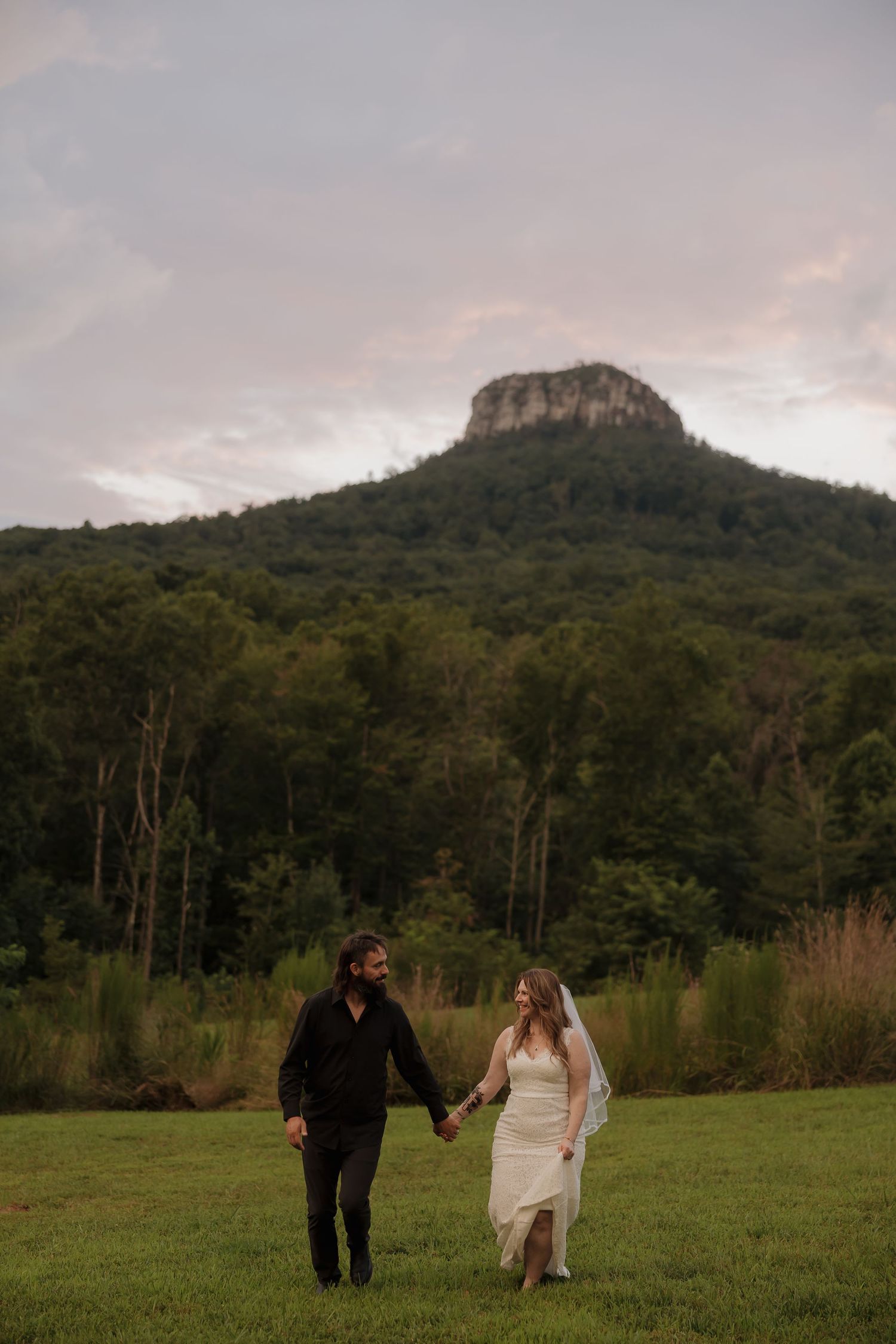 A couple in wedding attire walks hand in hand across a grassy field with a majestic mountain peak in the background.
