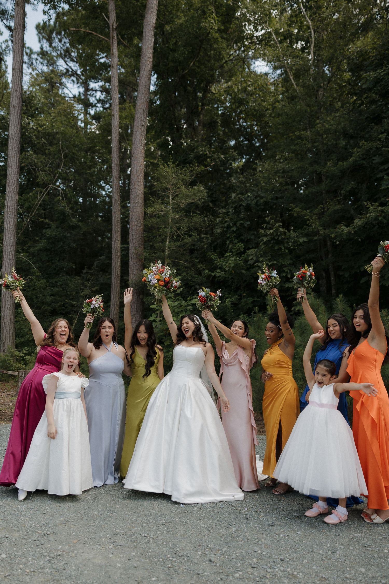 Bridal party in rainbow colored dresses holding bouquets against wooded backdrop.
